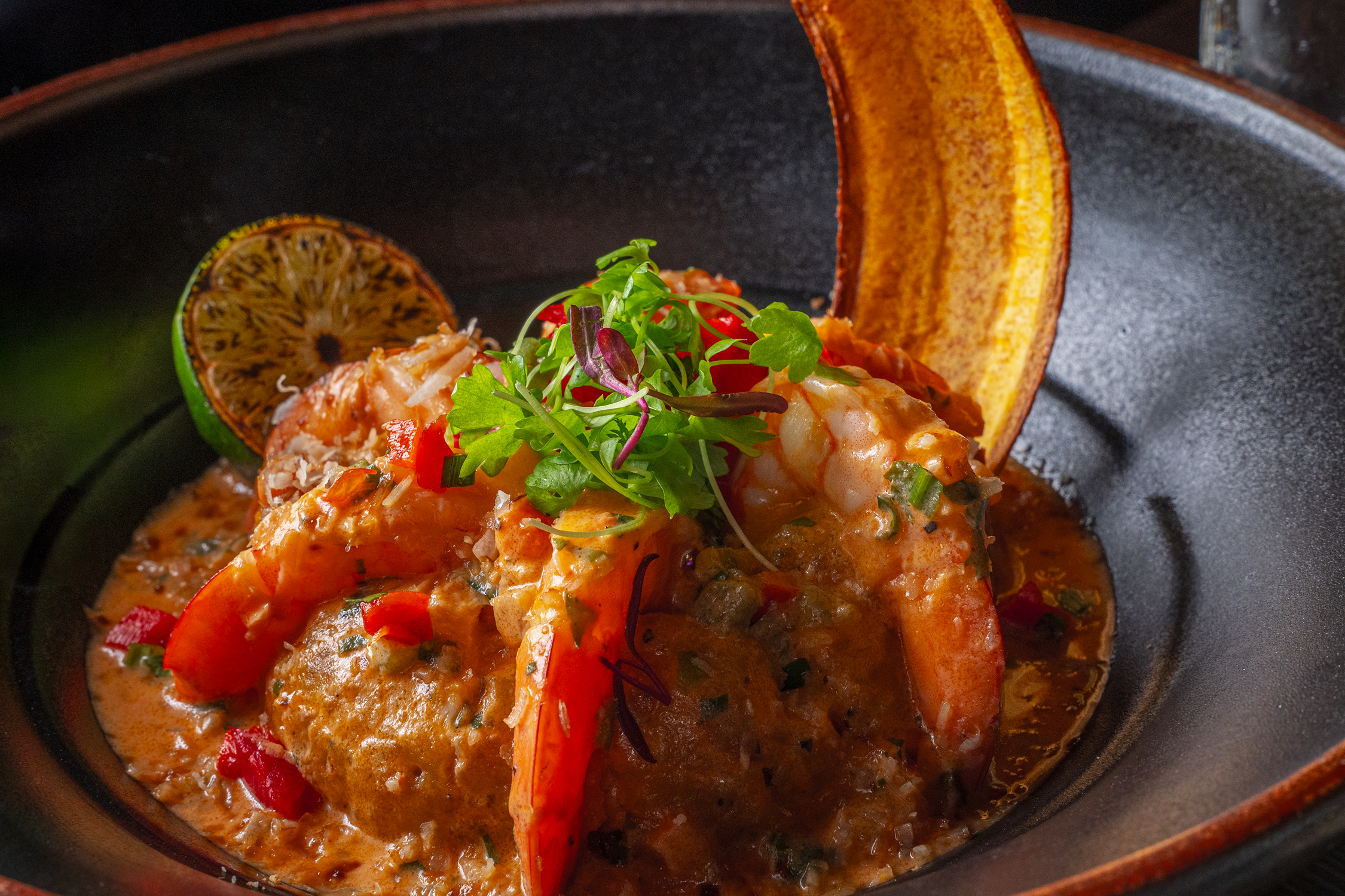 Close-up of a spicy seafood stew with shrimp, served with a lime wedge, corn tortilla chips, and garnished with microgreens.