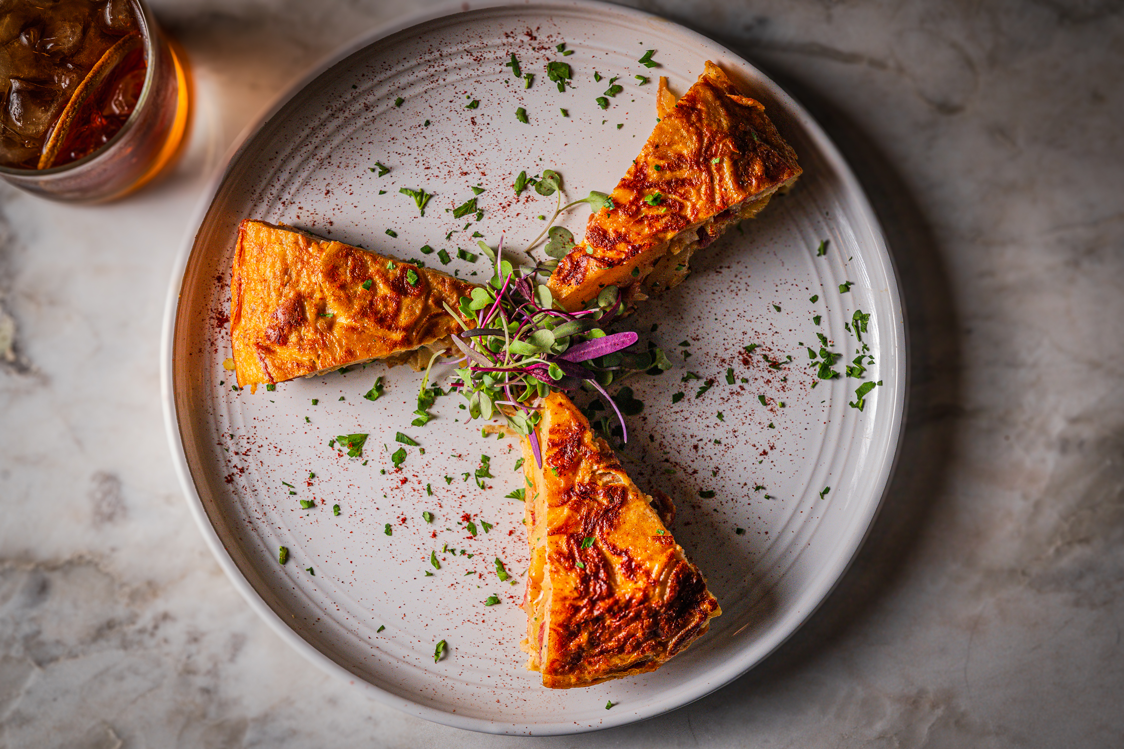 Three slices of quiche garnished with chopped herbs and microgreens on a white plate, with a glass of iced tea in the background.