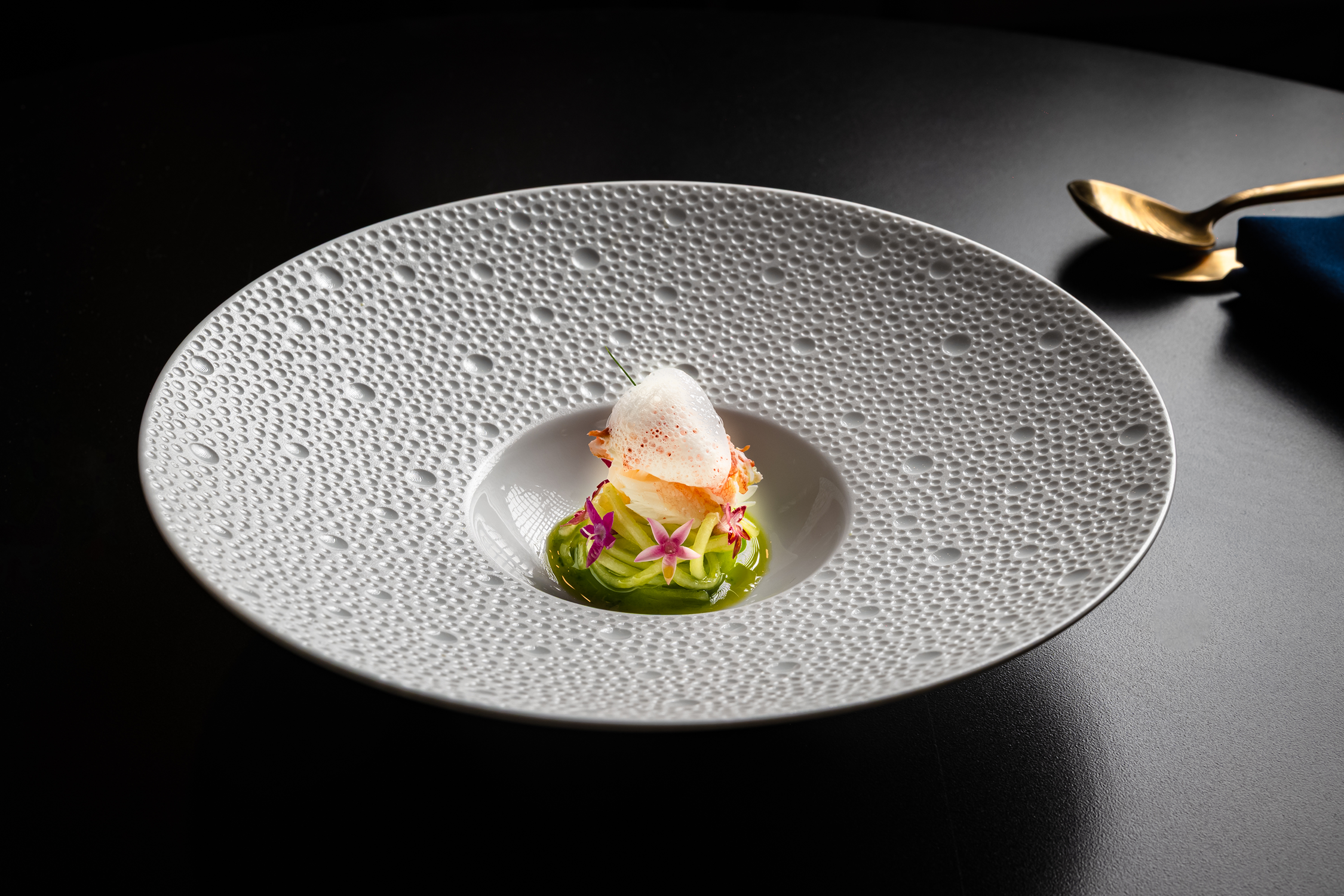 Elegant plated dish with a green vegetable base, topped with a frothy white foam and garnished with pink edible flowers on a textured white plate.