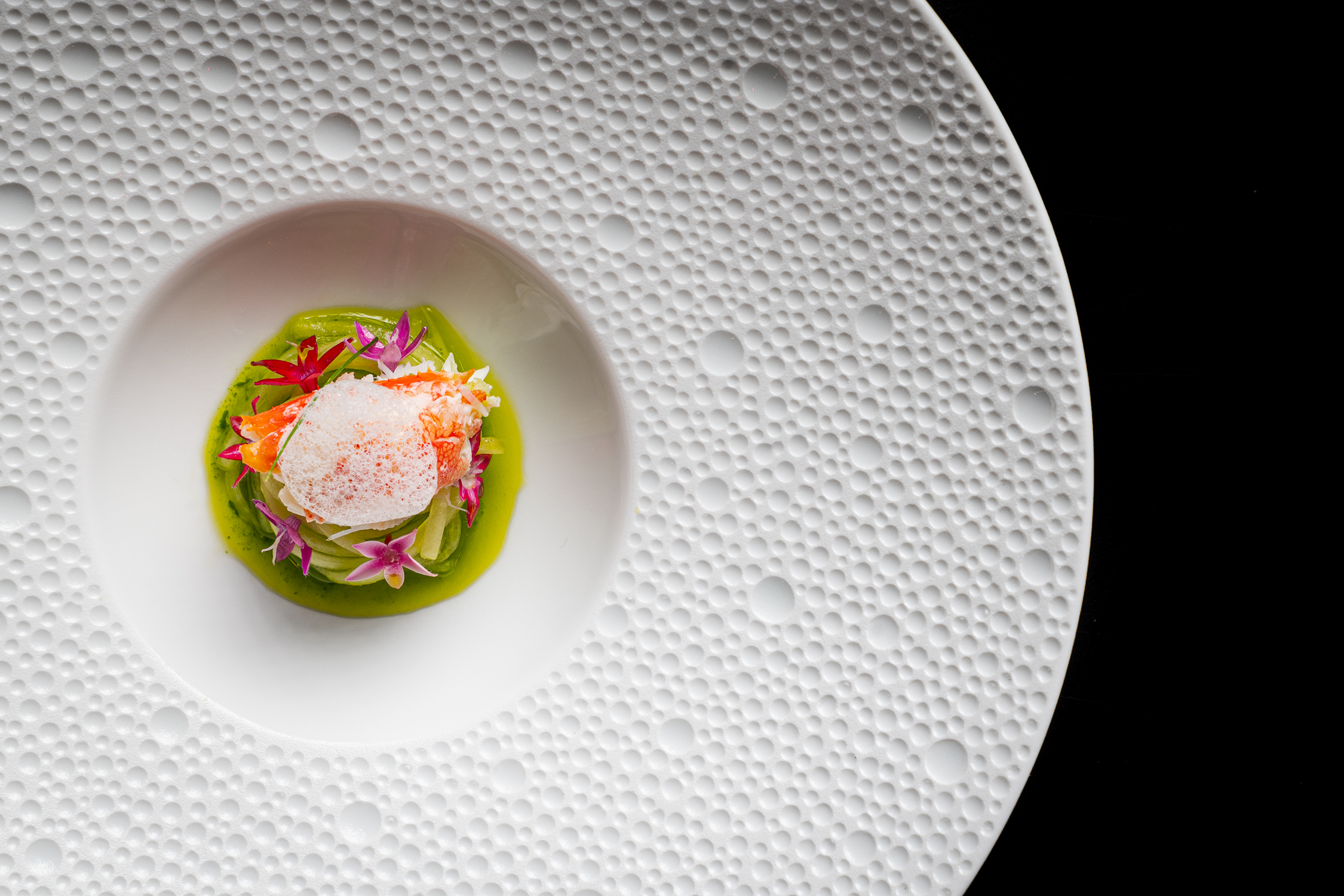 High-angle view of a gourmet seafood dish garnished with edible flowers on a textured white plate with a black background.