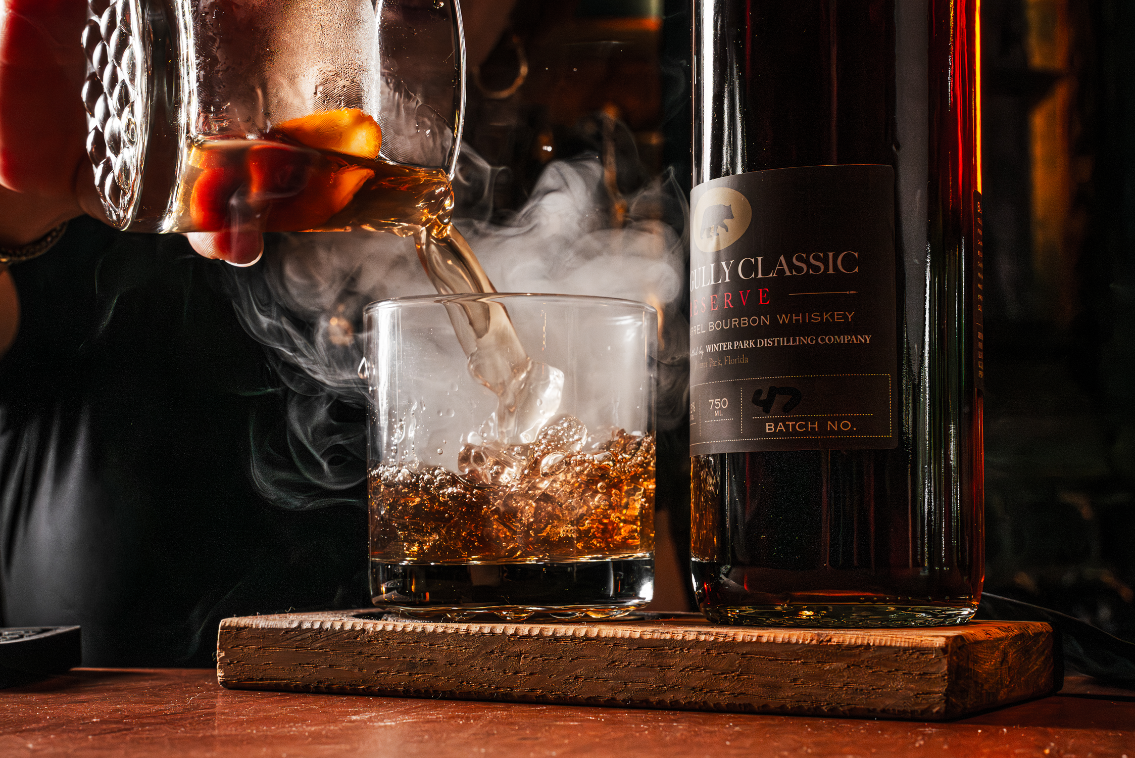 Pouring bourbon whiskey from a glass into a tumbler with ice, with the whiskey bottle nearby on a wooden surface.