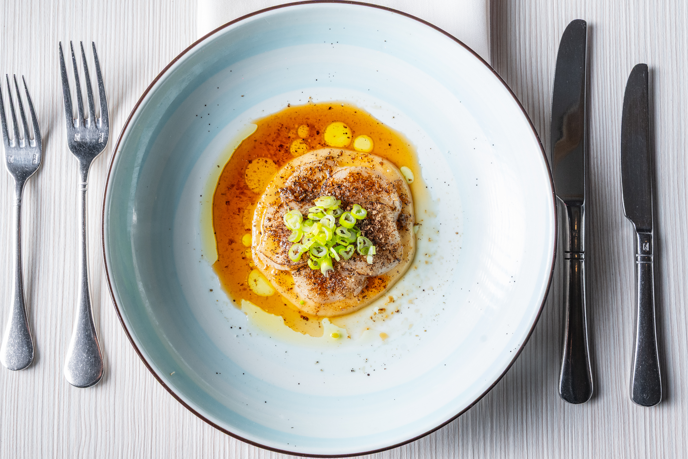 A plated dish featuring a cooked scallop topped with chopped green onions, surrounded by a sauce with a glossy appearance and garnished with black pepper.
