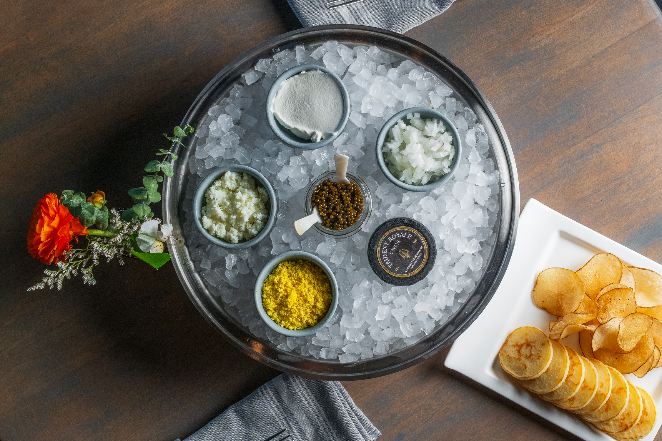 A tray with ice and small bowls containing various ingredients, surrounded by a small bouquet of orange, white, and green flowers, and a plate of potato chips on the side.
