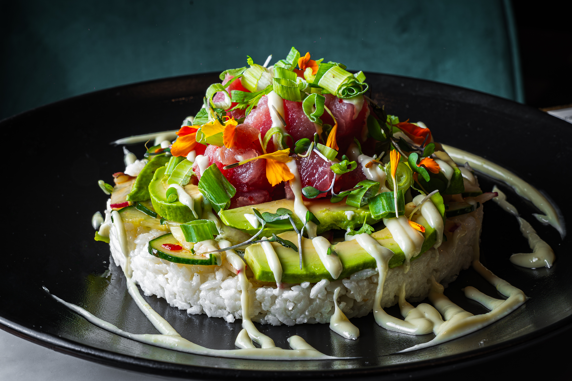 A plated dish featuring rice, sliced avocado, chopped green onions, diced raw fish, and garnished with edible flowers drizzled with white sauce, served on a black plate.
