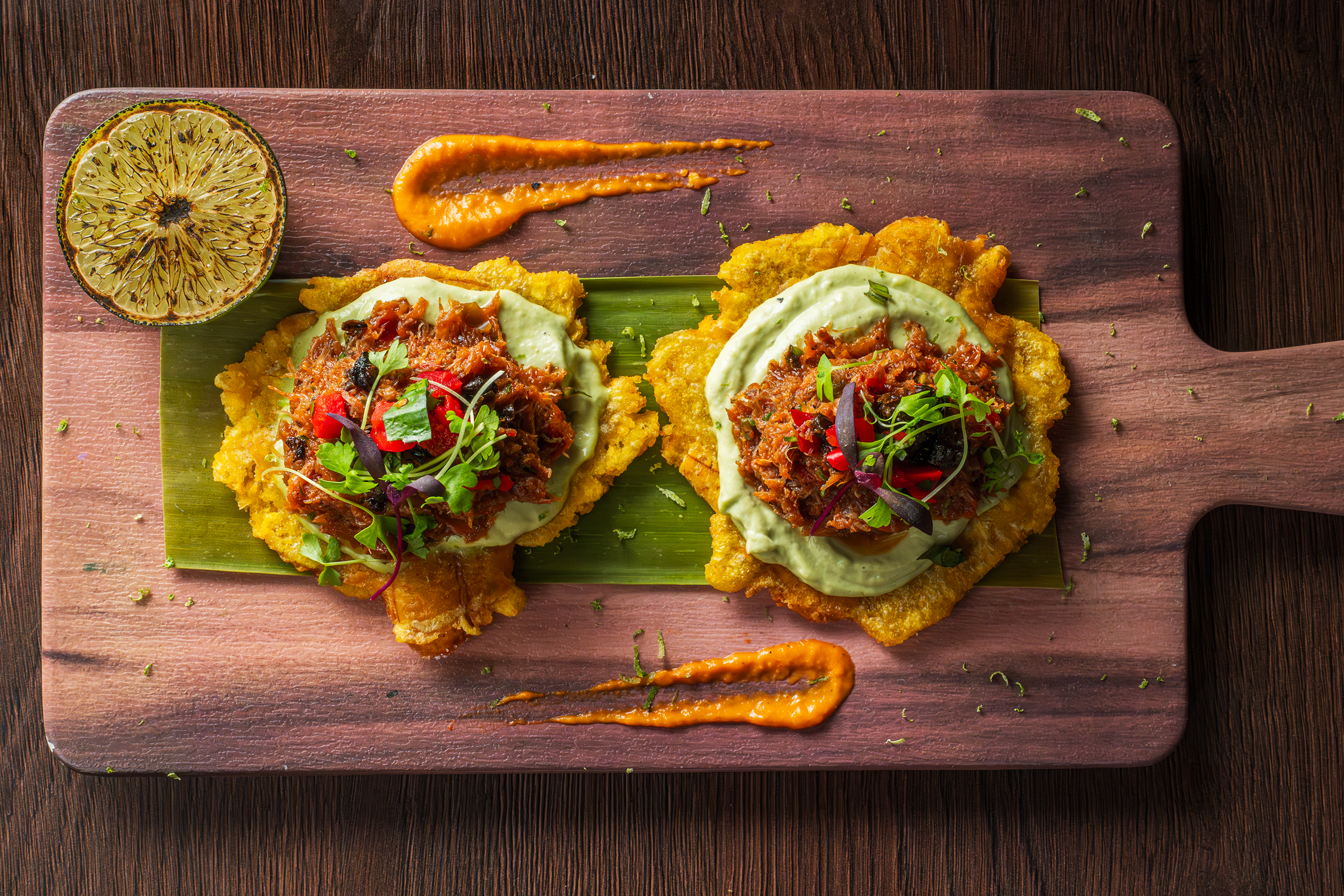 Two pieces of fried fish topped with a mixture of shredded meat, chopped vegetables, and herbs, served on a wooden board with a lime half and dollops of orange sauce.