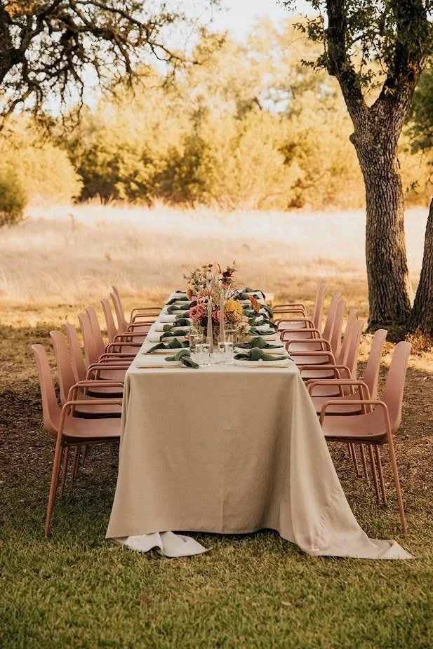 Long outdoor dining table with a cream tablecloth, pink chairs, floral centerpiece, and place settings beneath oak trees.