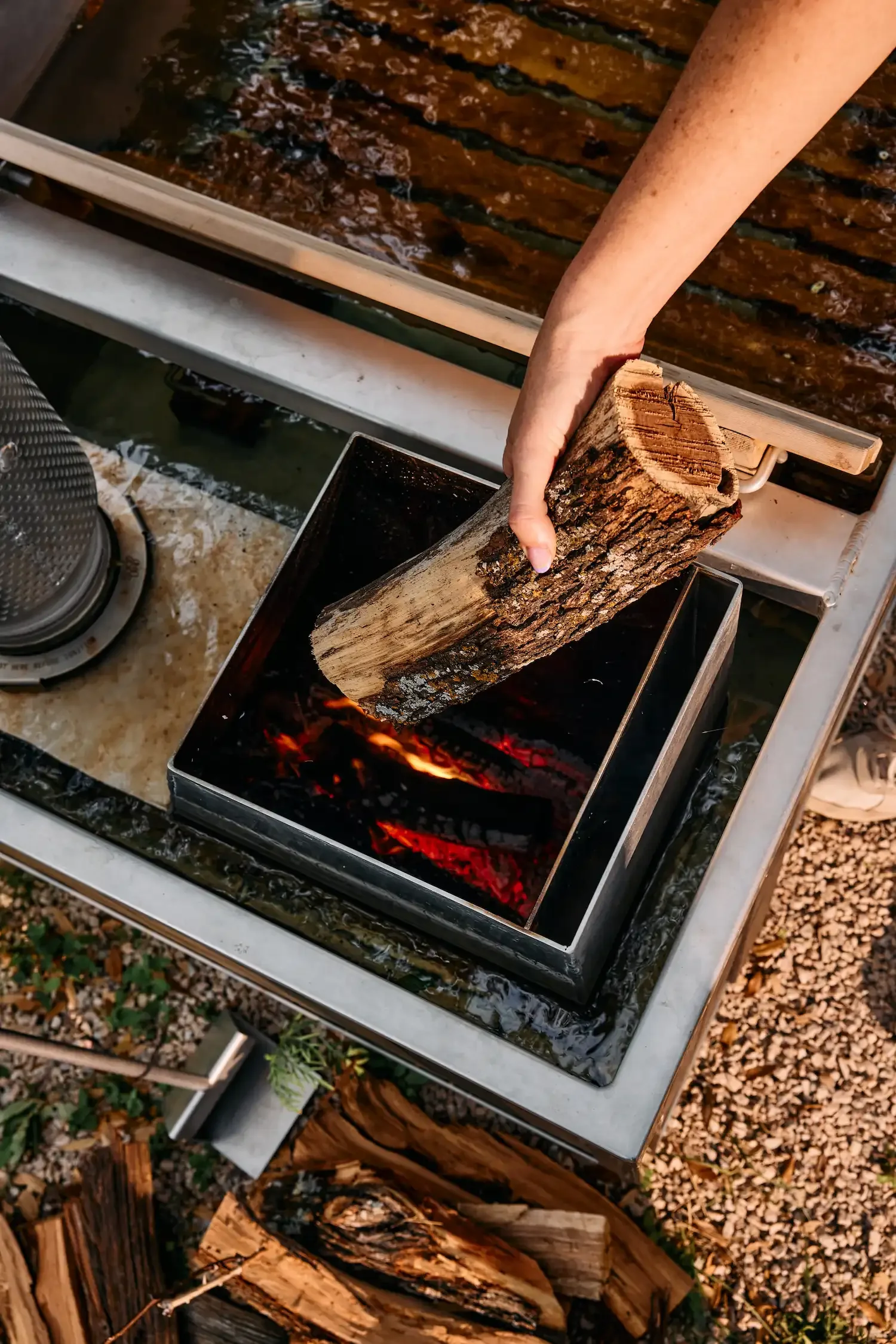 Hand placing a log into the glowing firebox of a wood-fired hot tub, with stacked firewood on the gravel below