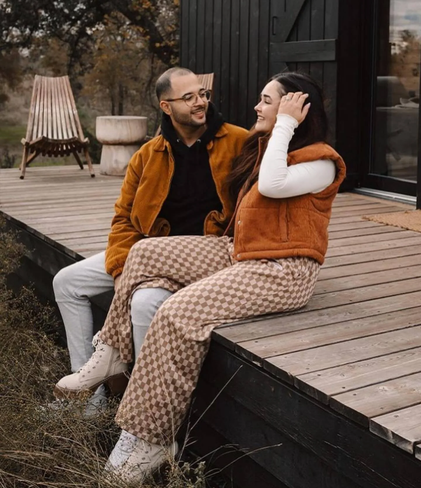 A couple sits together on a wooden cabin deck, wearing casual clothing beside an outdoor chair.
