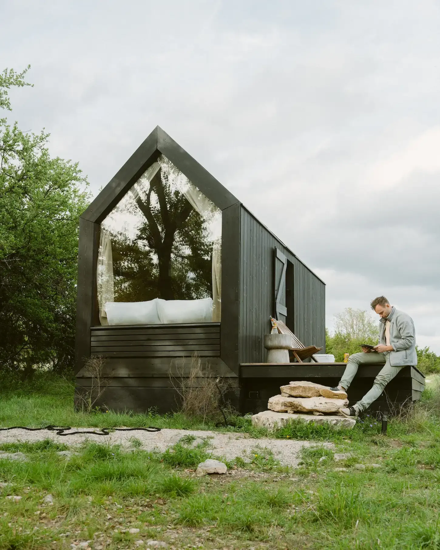 Guest reading a tablet on the edge of a deck beside a black A-frame cabin with a large glass window and a bed inside
