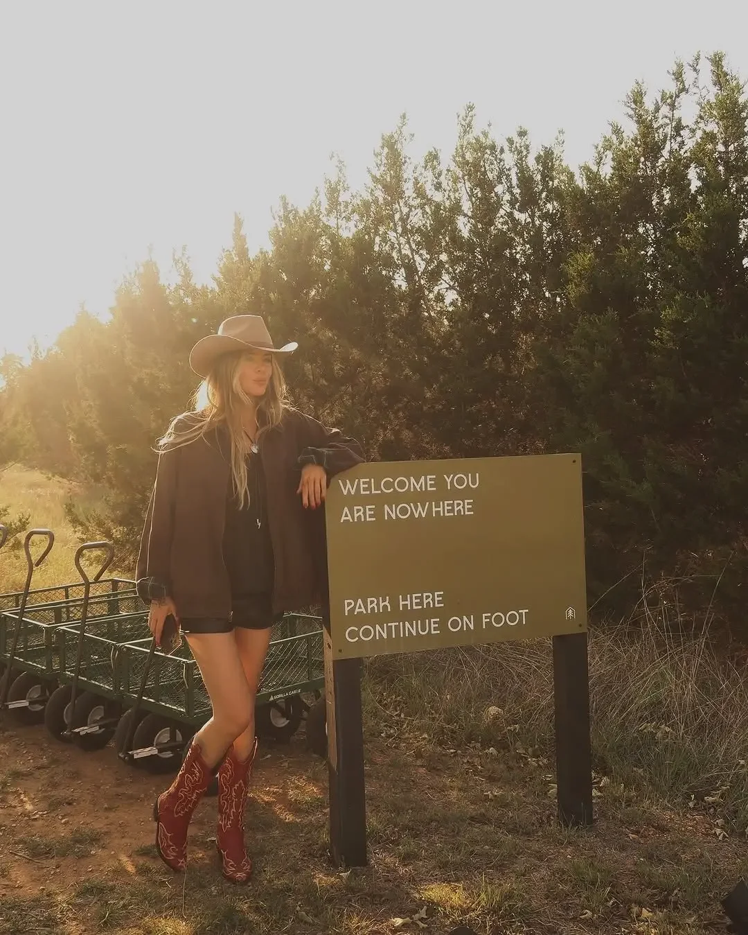 A guest wearing a cowboy hat and red boots stands beside a NowHere welcome sign near several luggage carts.
