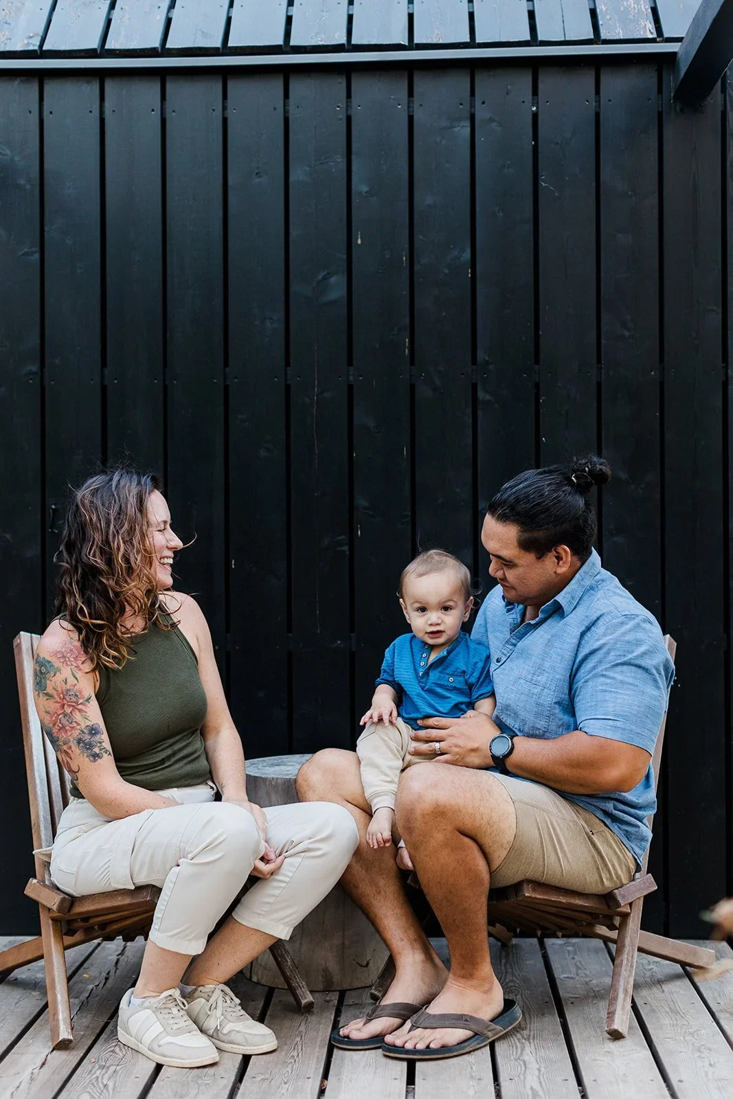 Erika's family seated in wooden chairs on a deck, with a baby on a man's lap, in front of a black wood wall