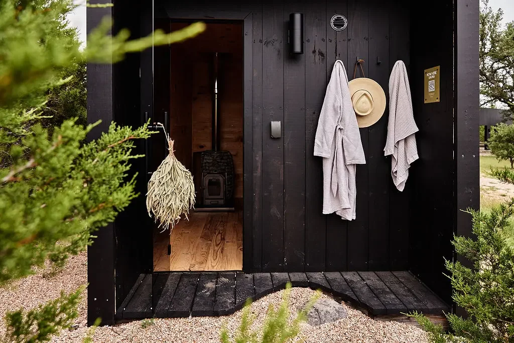 Black wood sauna entrance with an open door, a stove inside, and robes and a straw hat hanging on the wall outside