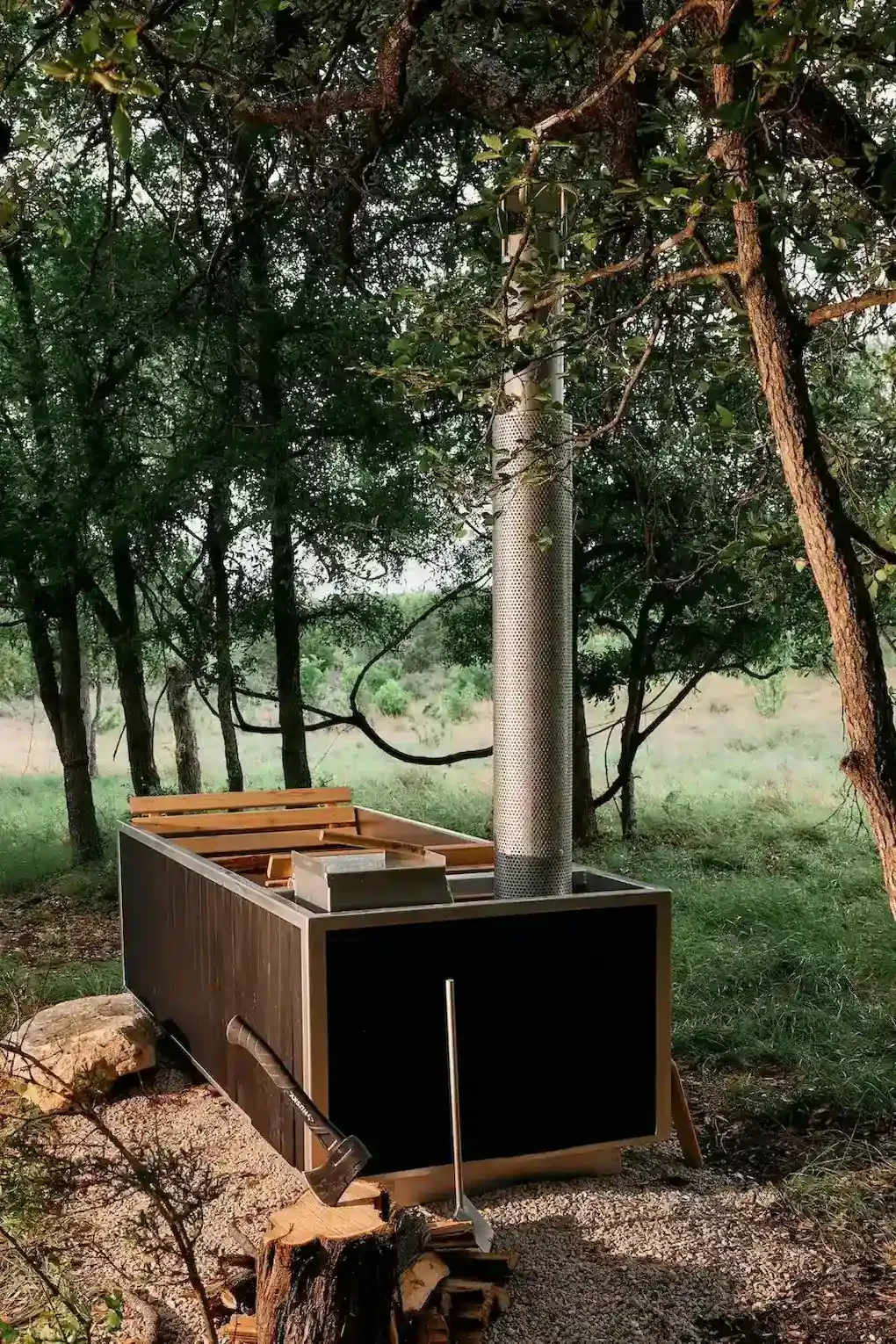 Wood-fired hot tub with a metal chimney among oak trees, with an axe in a stump and stacked firewood in front