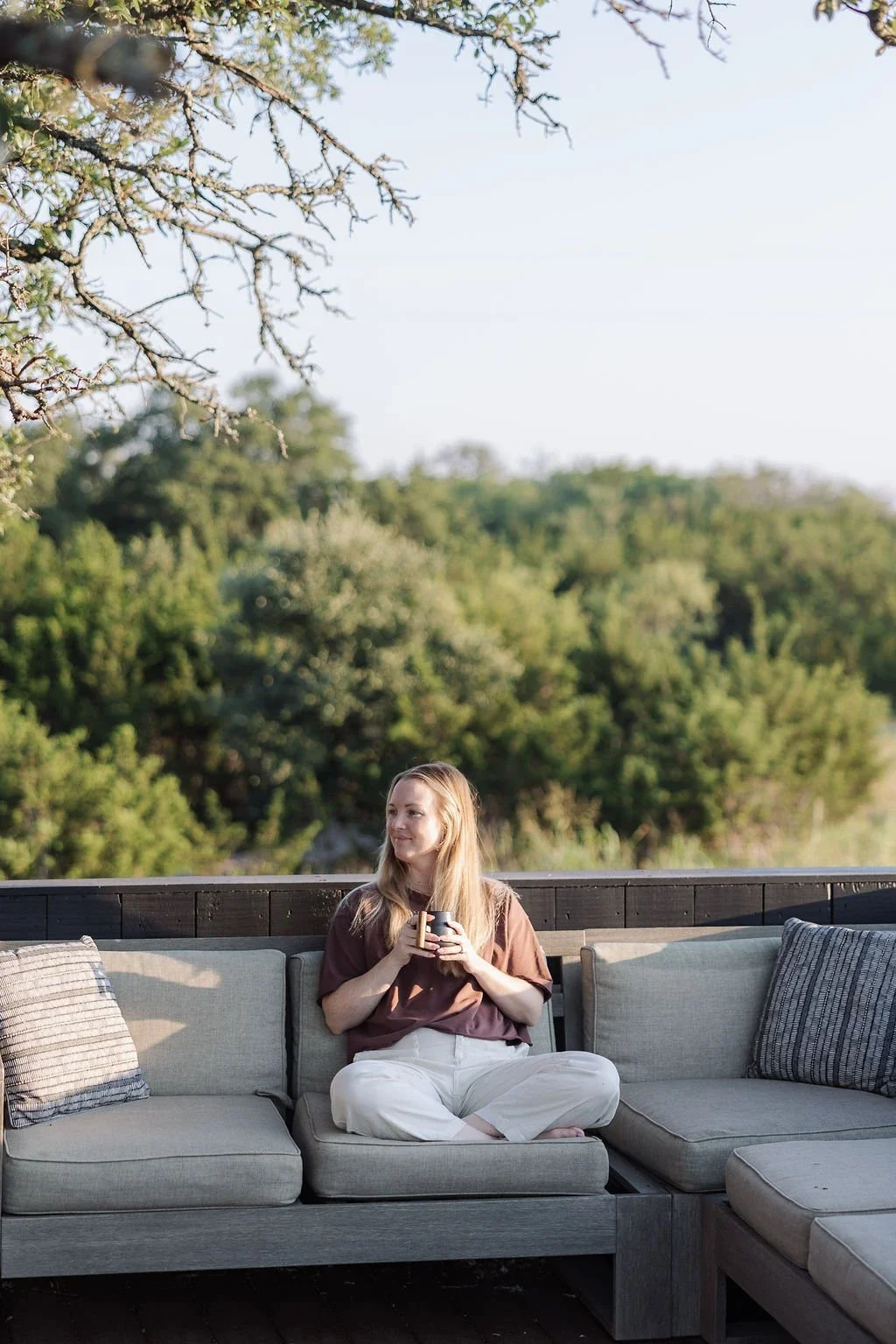 A guest sits cross-legged on an outdoor sofa holding a mug, surrounded by trees and greenery.