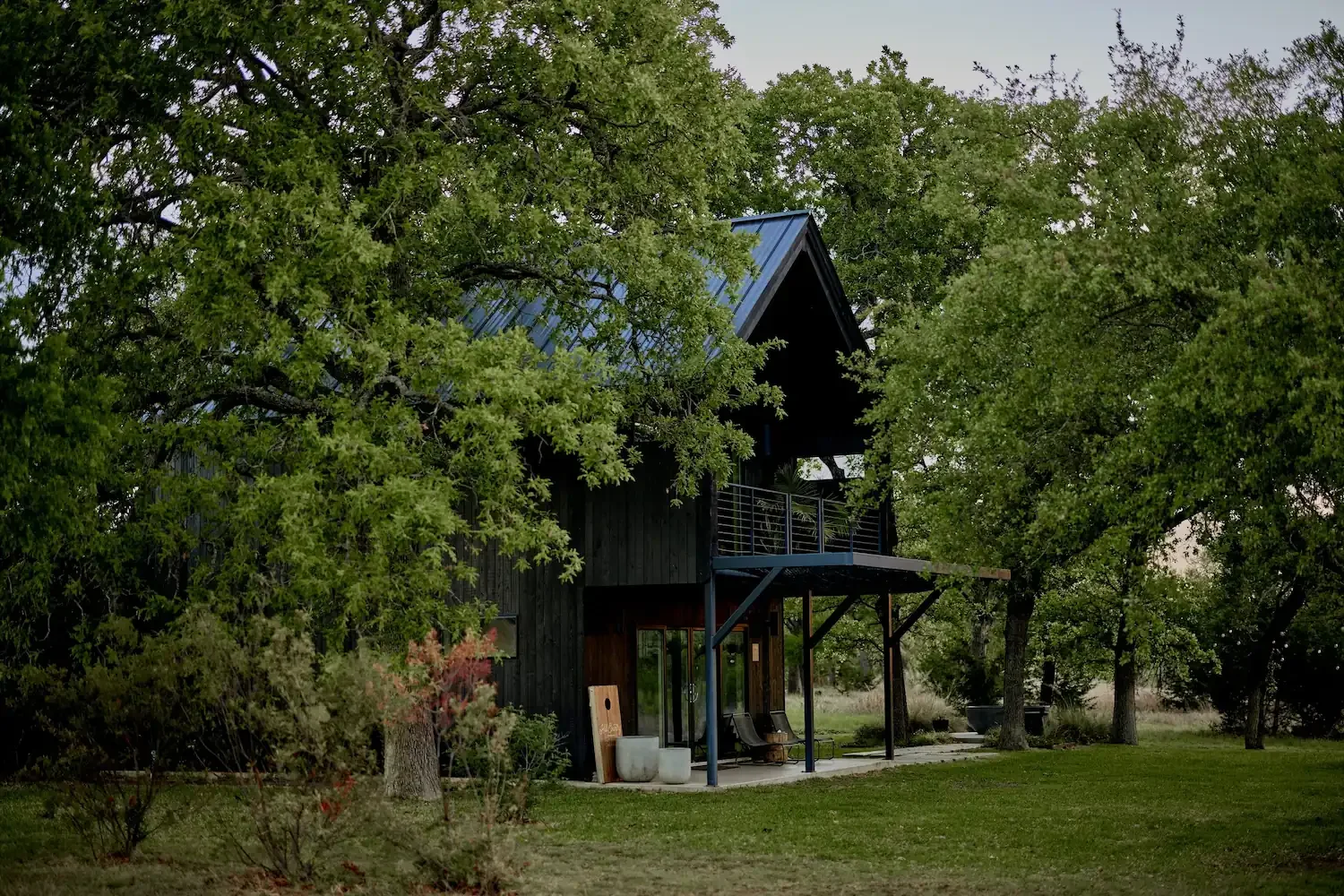 A black cabin with a metal roof, upper balcony, and covered patio surrounded by trees at NowHere.