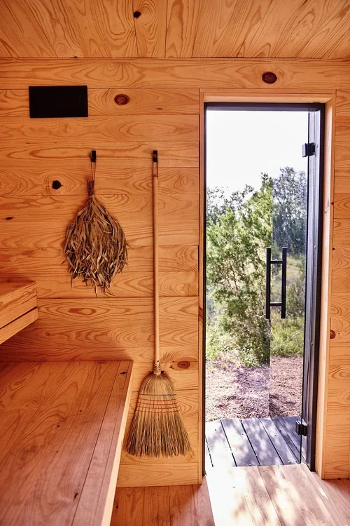 Wooden sauna interior with a bench, hanging bundle of branches, broom, and glass door overlooking trees.