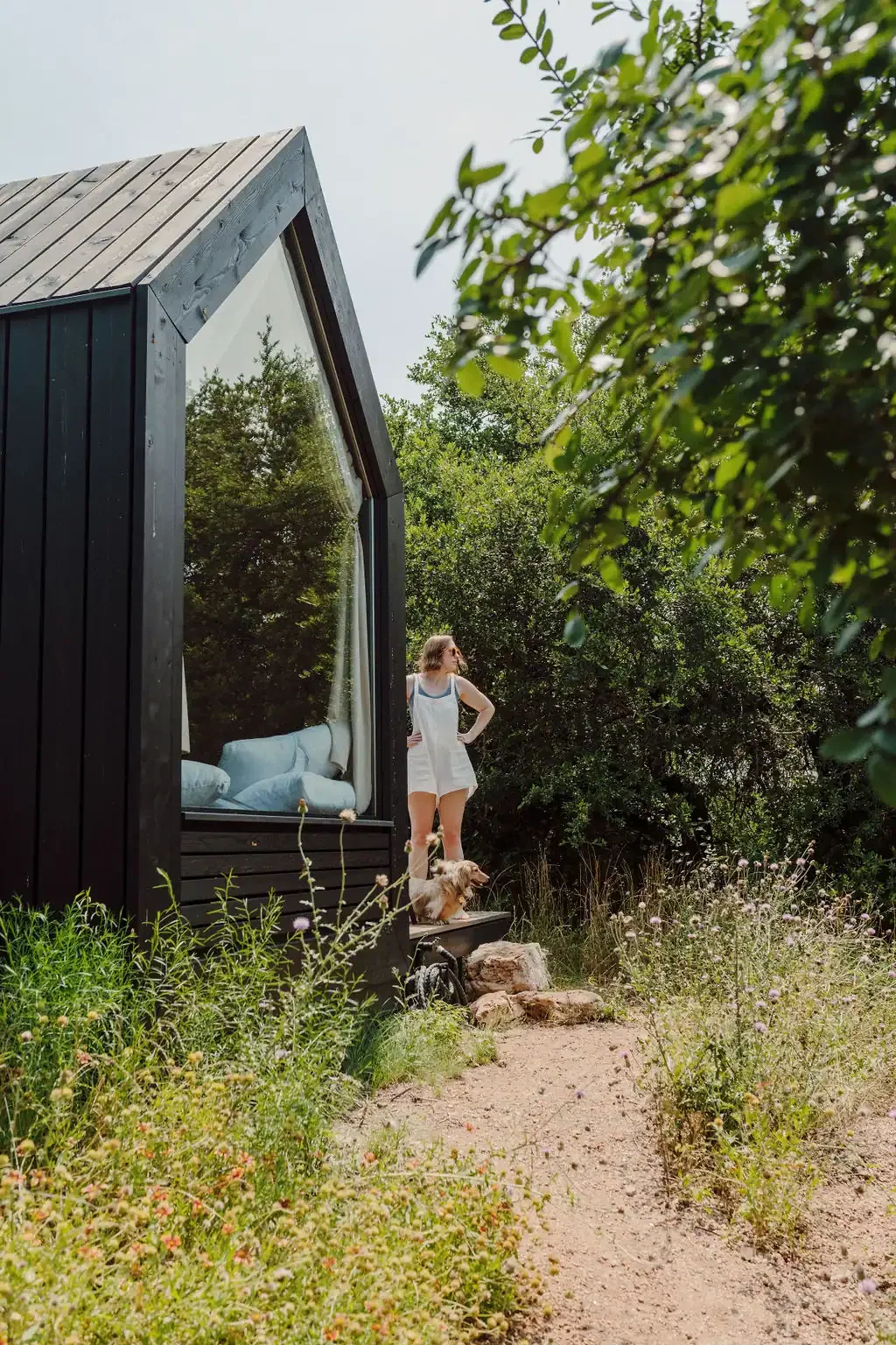 A guest stands beside the Sage cabin with a small dog near the large triangular window, surrounded by trees and wildflowers.