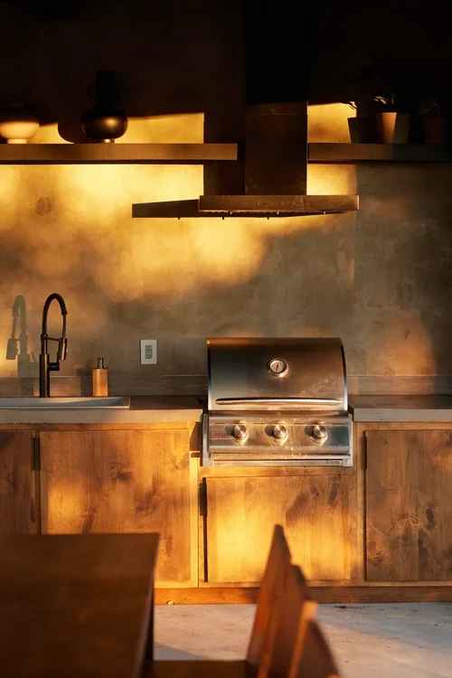 Outdoor kitchen with wooden cabinets, a stainless steel grill, a sink, and a black range hood.