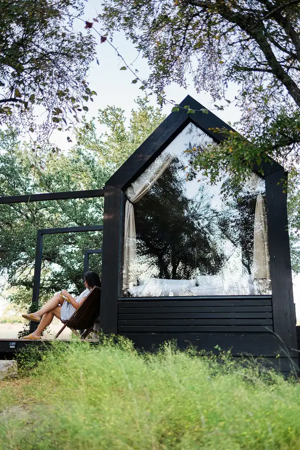 Guest reading in a wooden lounge chair beside Agave, a black A-frame cabin with a large glass window, under oak trees