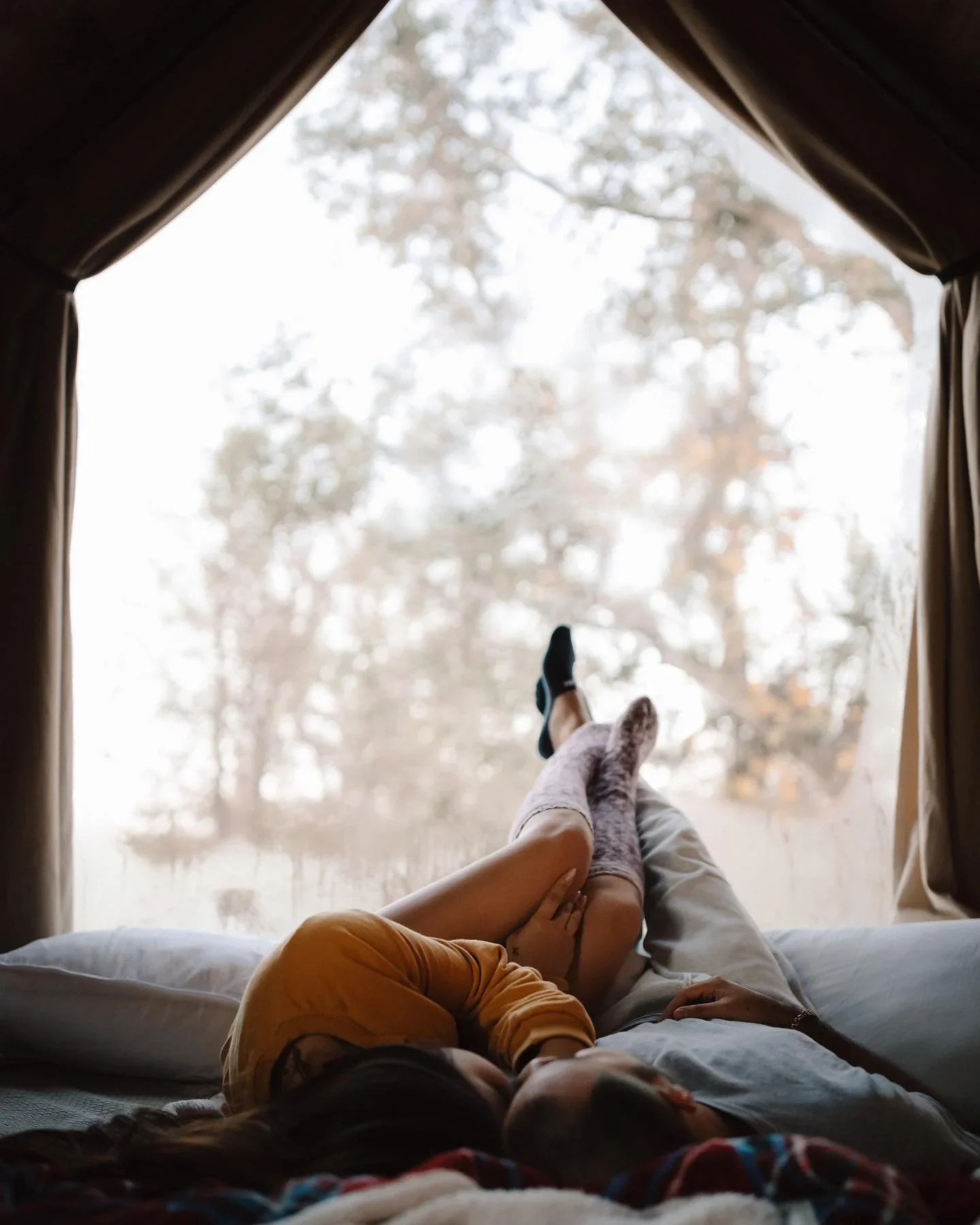Couple lying on a bed with their legs crossed, framed by open curtains overlooking trees.