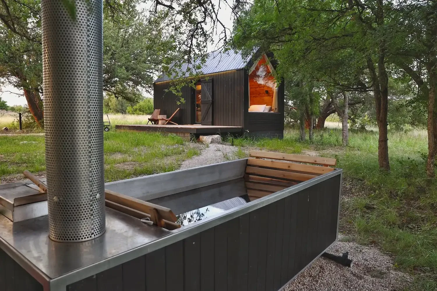 An outdoor soaking tub with a metal chimney sits in front of a black cabin surrounded by trees.