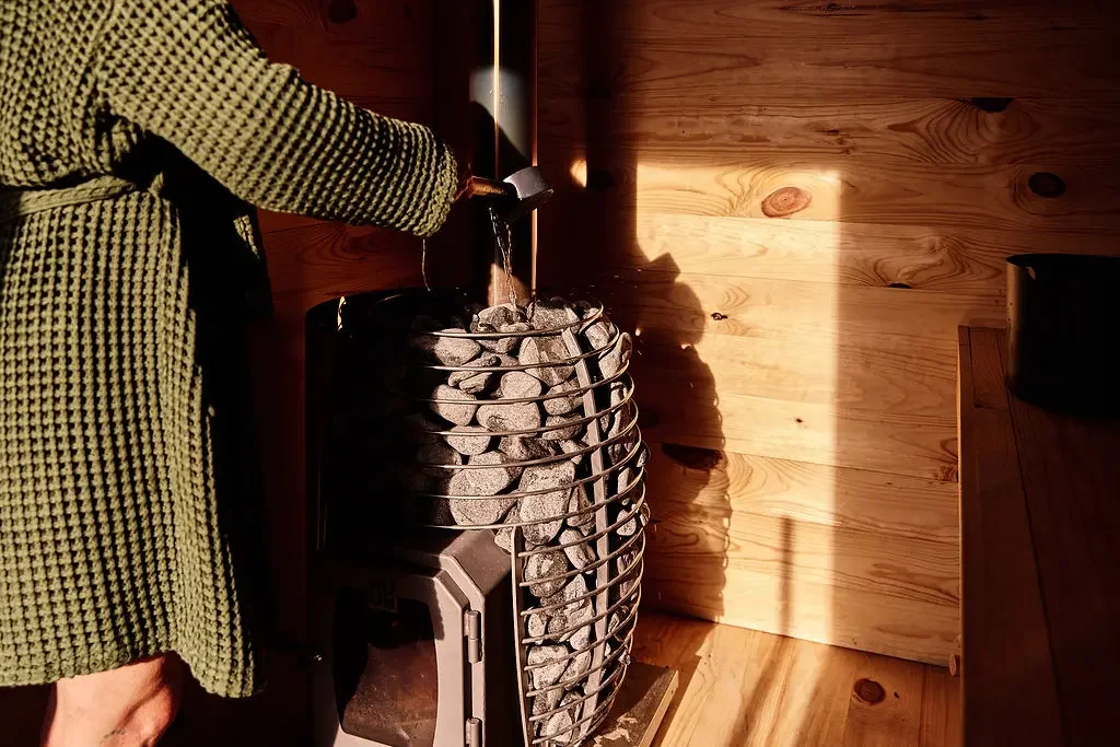 Guest in a green waffle-knit robe pouring water from a ladle onto the stones of a wood-fired sauna stove