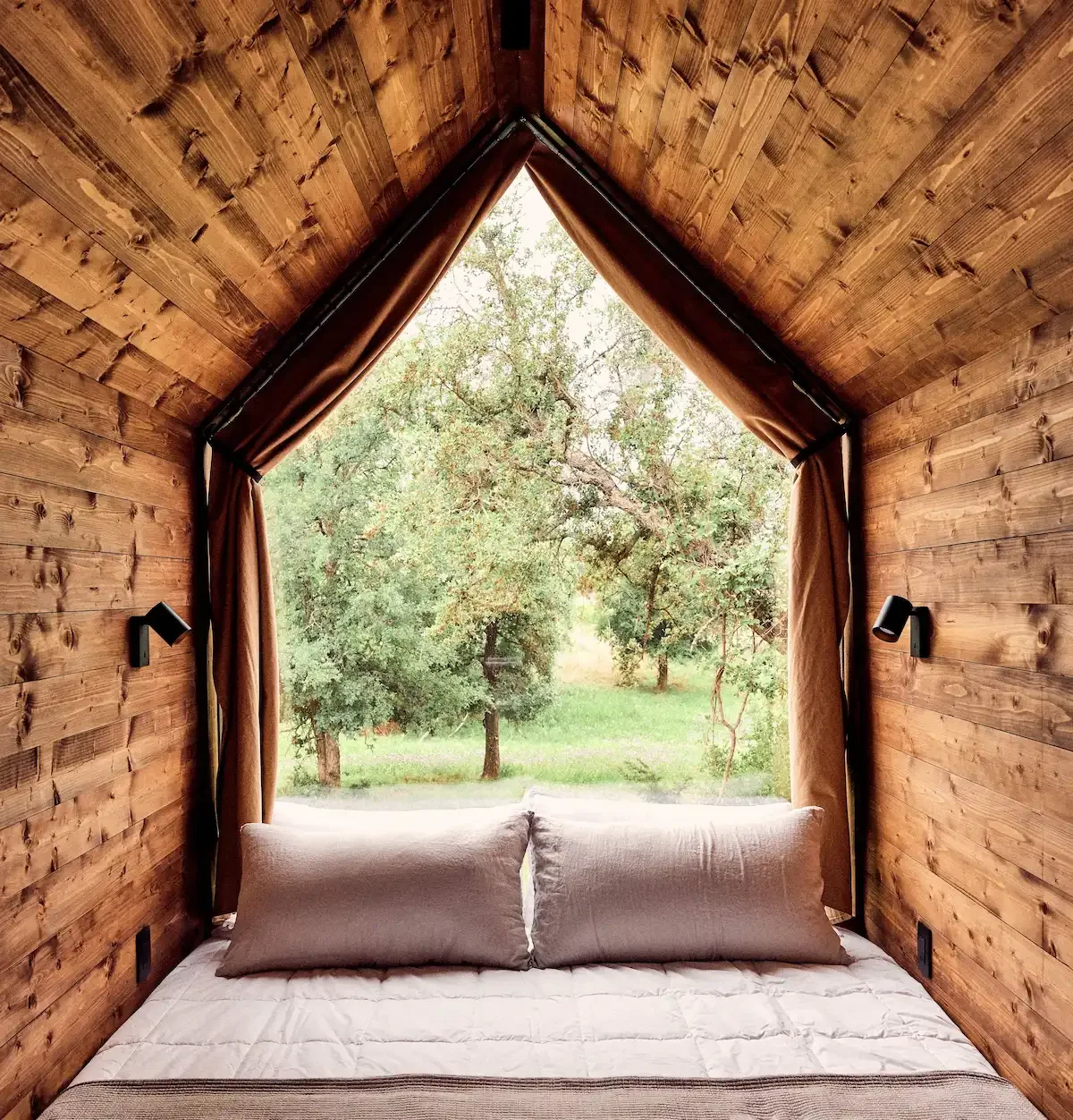 A bed with two pillows in a wood-paneled room with a triangular window overlooking trees.
