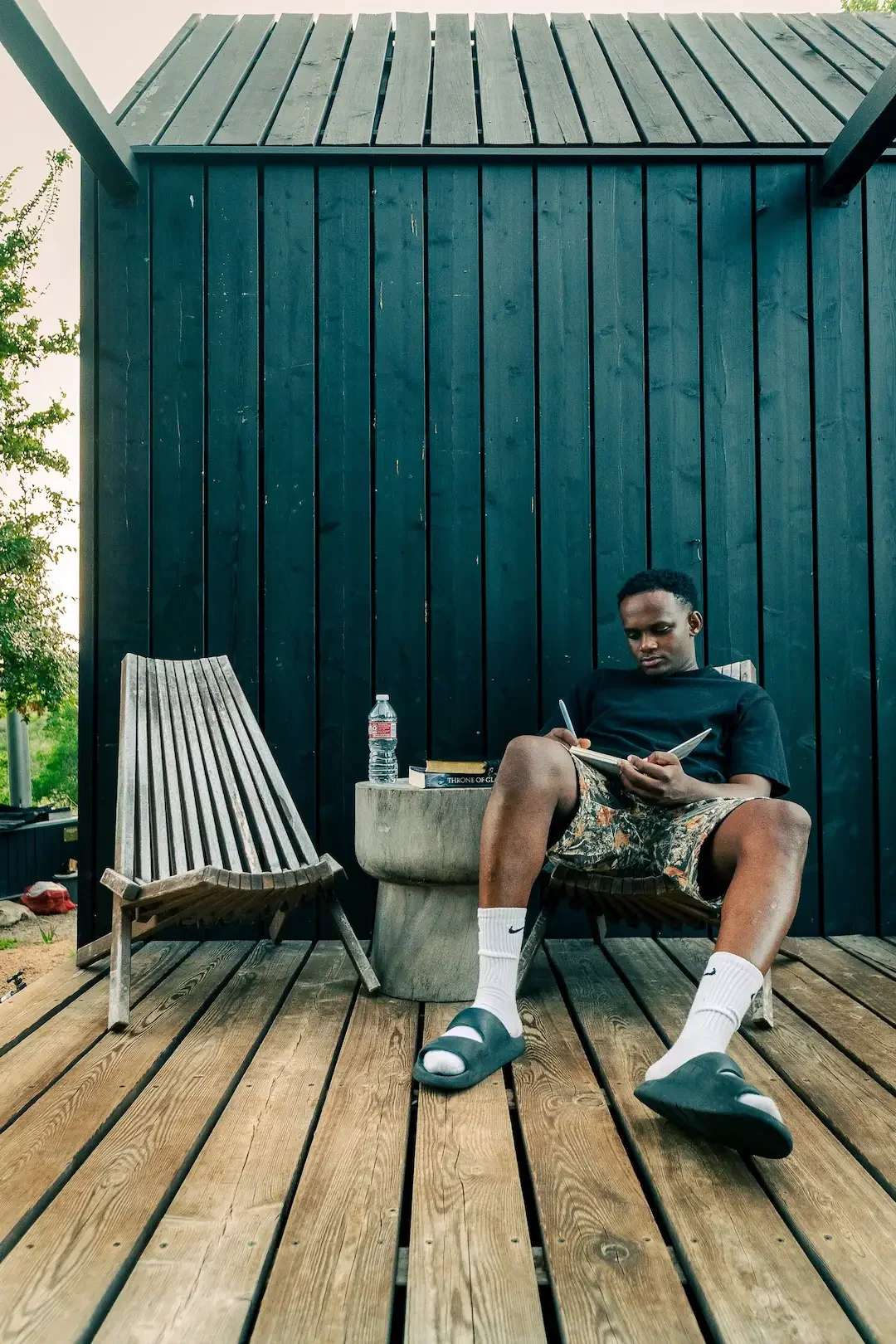 A guest writes in a notebook while seated on a wooden deck beside an empty chair.