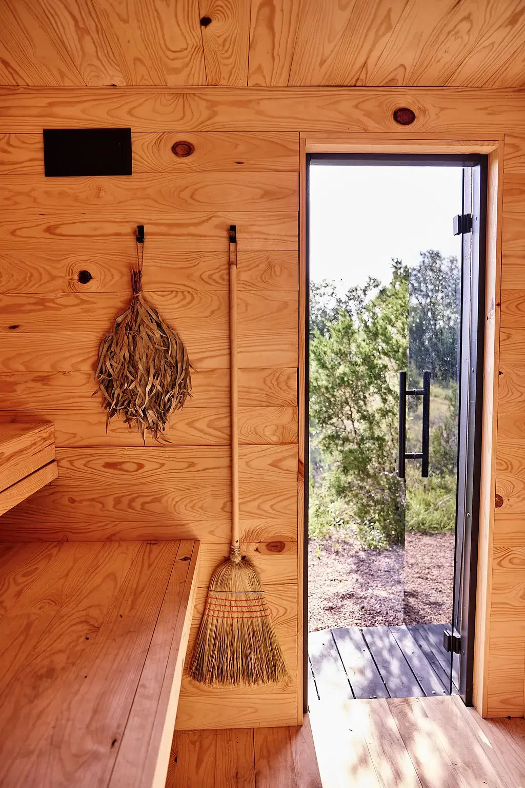 Pine-paneled sauna interior with benches, a hanging leaf bundle and a broom, and an open glass door facing trees