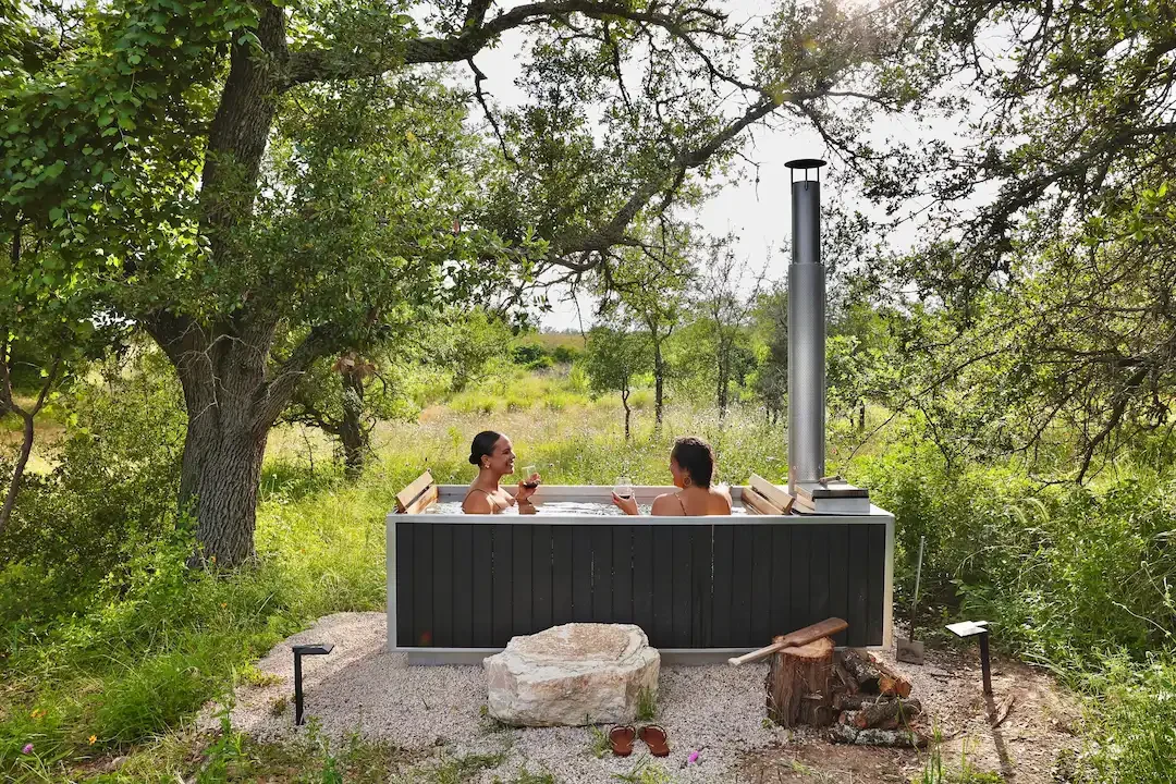 Two guests soaking in a wood-fired hot tub with glasses of red wine, surrounded by oak trees and a grassy meadow