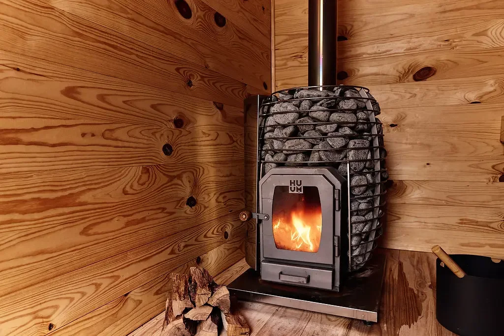 Wood-burning sauna stove with a lit fire and a cage of stones, in a pine-paneled corner with split firewood beside it
