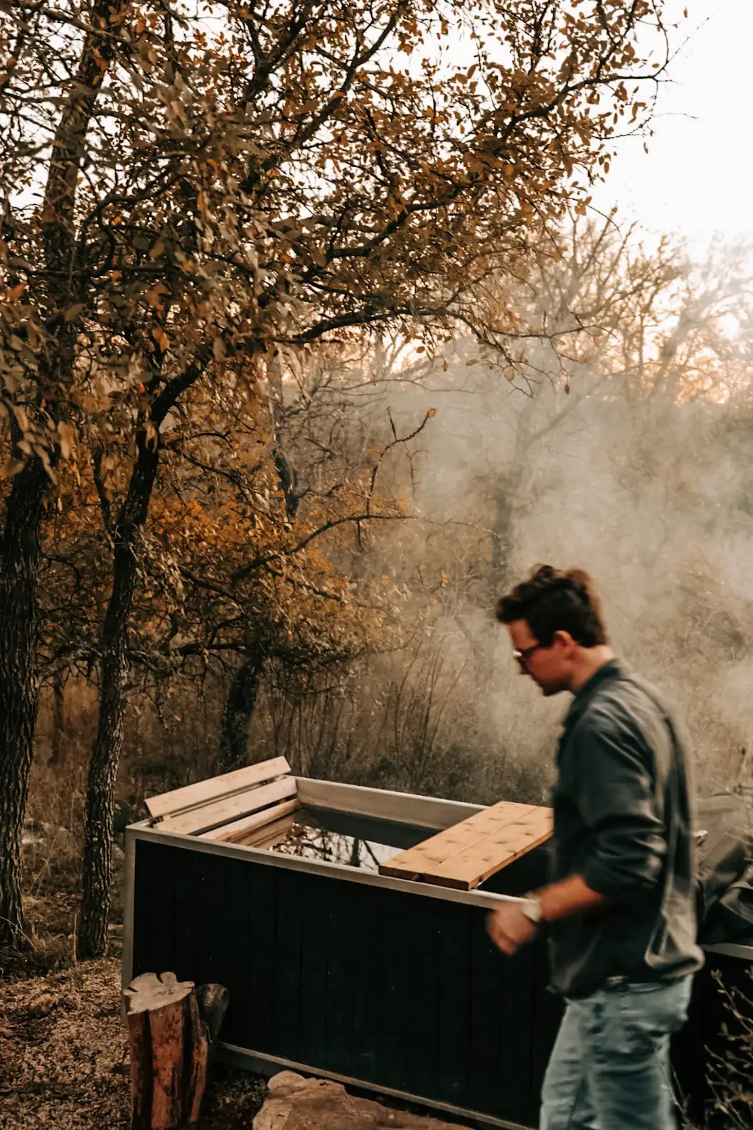 Guest in glasses and a gray jacket lifts a wooden lid on a hot tub, with smoke drifting through the trees behind