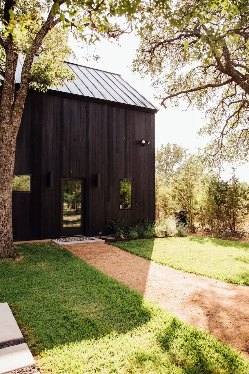 A black cabin with a metal roof sits among trees beside a grassy lawn and dirt path.