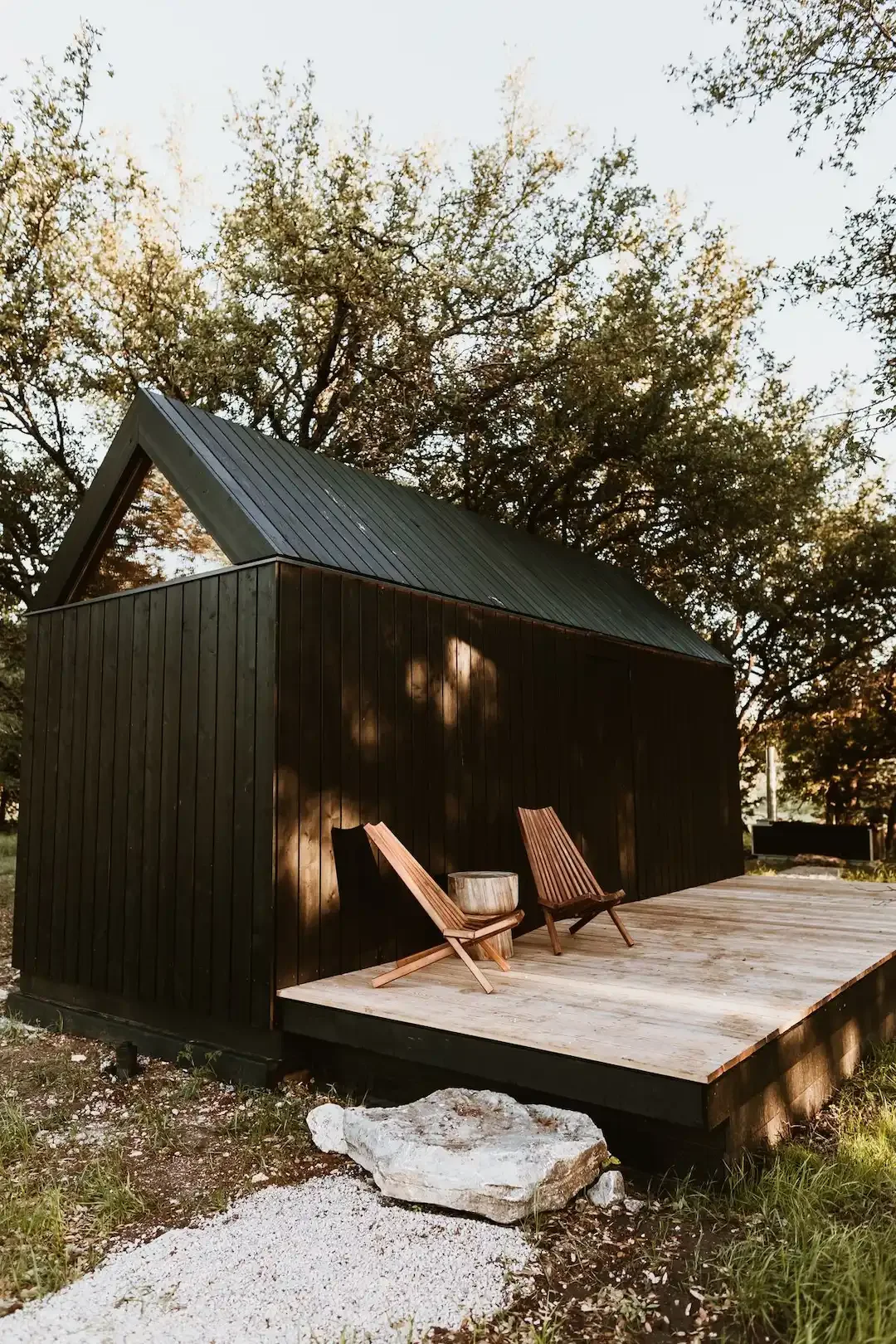 A black A-frame cabin with a wooden deck, two lounge chairs, and trees surrounding the structure.