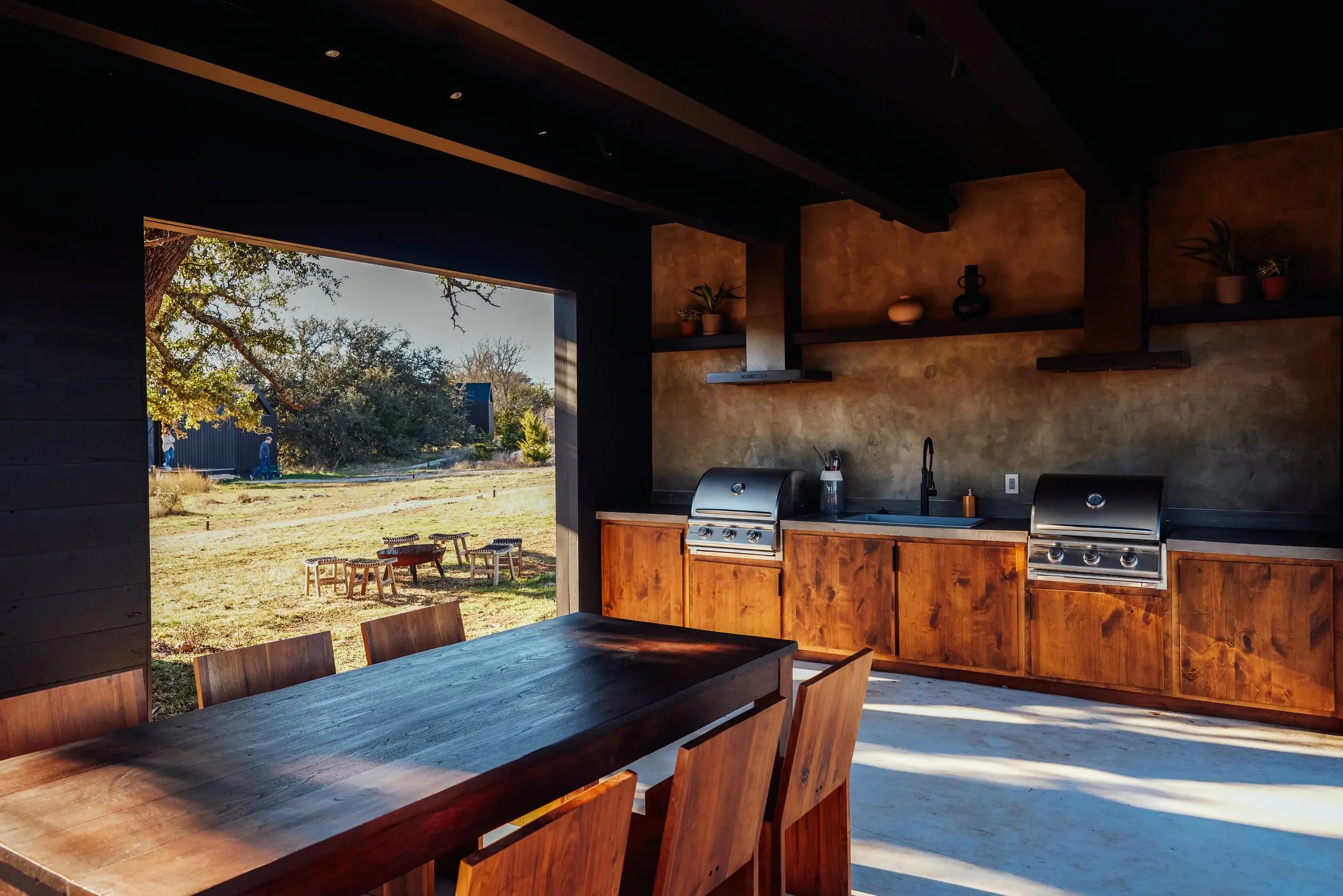 An outdoor kitchen with grills, wooden cabinets, and a large dining table overlooking the lawn.
