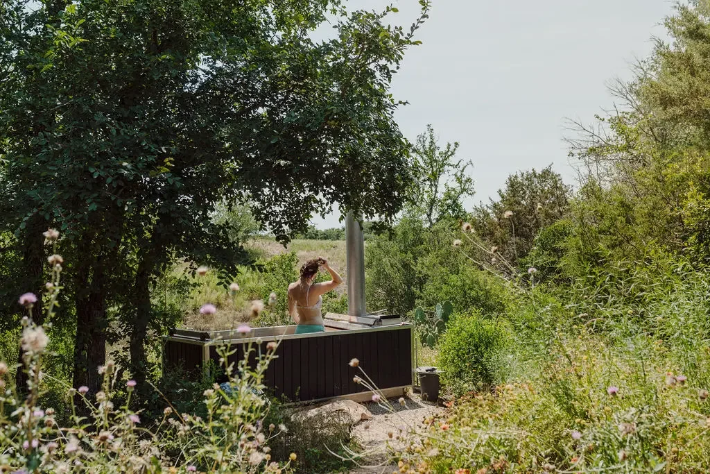 Guest in a bikini top standing in a wood-fired hot tub with a metal chimney, surrounded by trees and wildflowers
