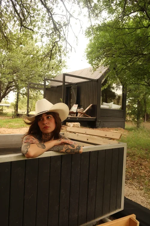 Woman wearing a cowboy hat rests her arms on the edge of a wooden hot tub with a cabin among trees behind her.