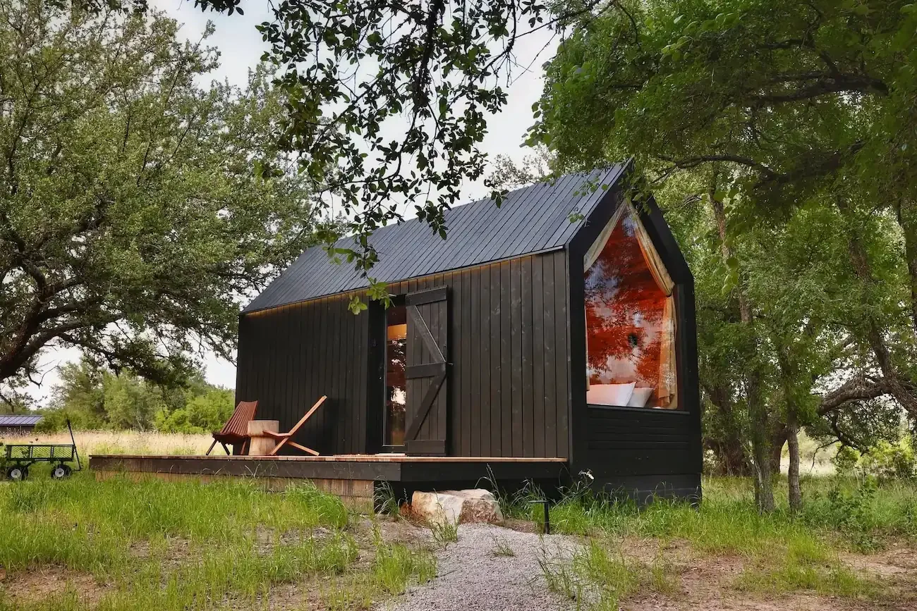 A black cabin with a metal roof, wooden deck, and two chairs sits among trees and tall grass.