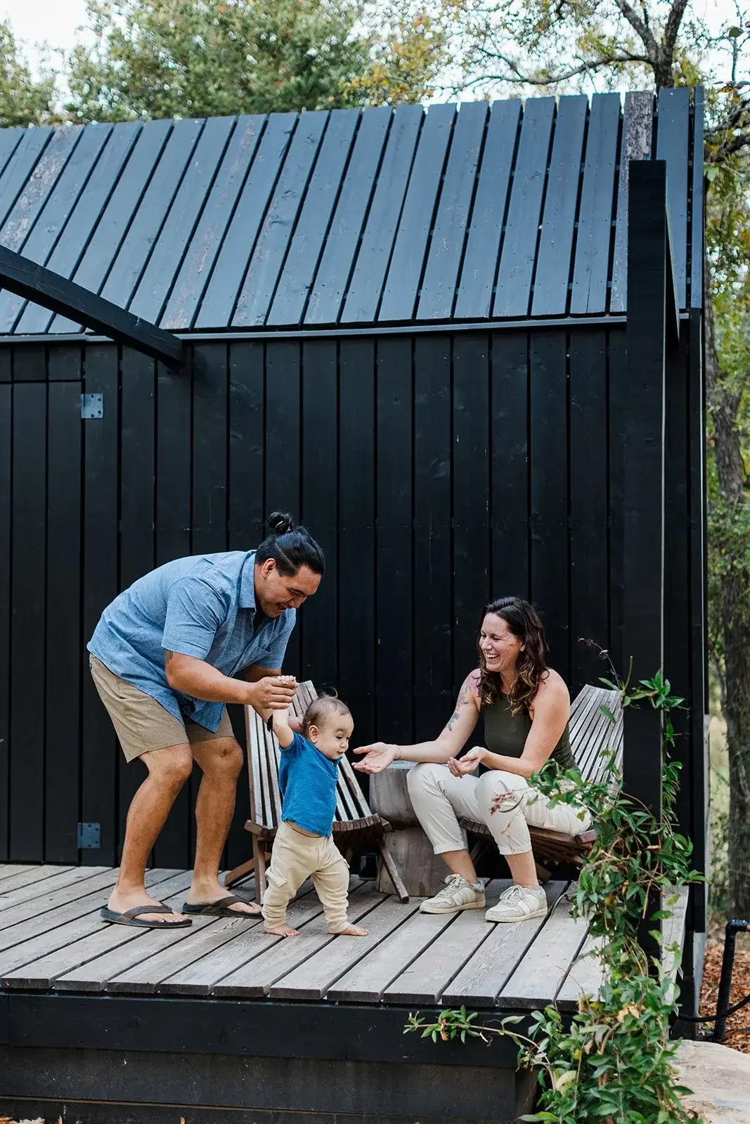 Erika's family on a wood deck outside a black cabin, a man helping a toddler walk while a woman sits nearby with open hands