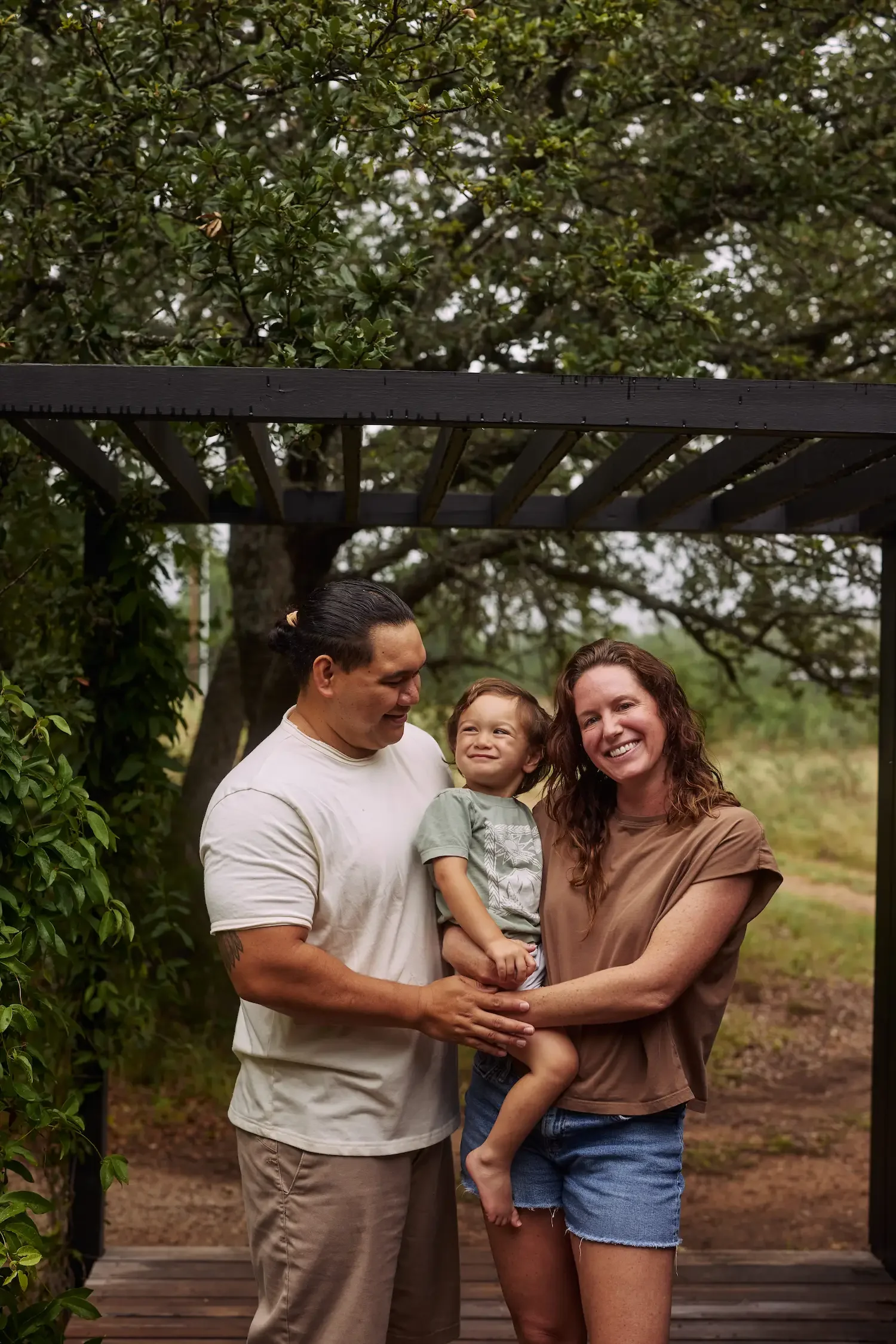 Kamalu and Erika standing together holding their child, under a black wood pergola with oak trees and a field behind