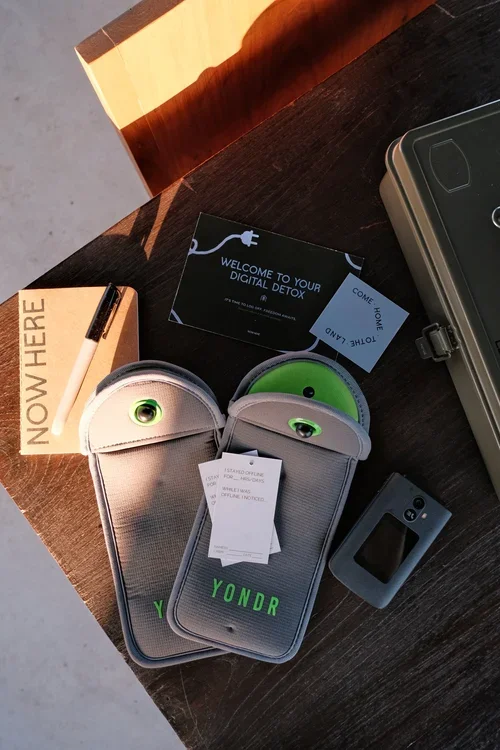 Yondr phone pouches, a digital detox welcome card, and a phone pouch lock sit on a wooden table beside a NowHere notebook.