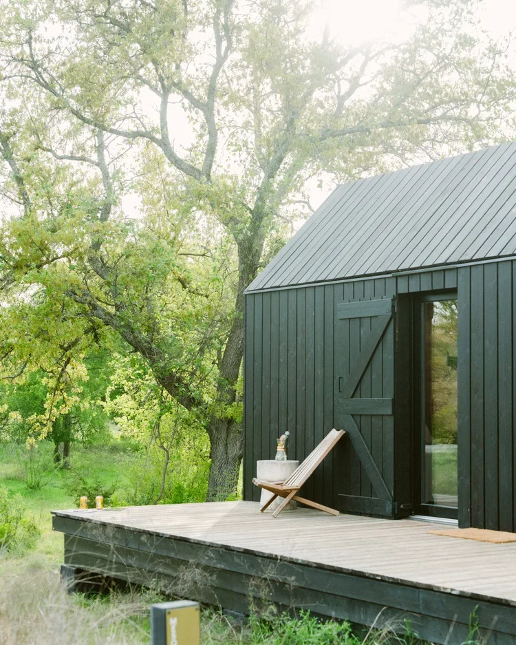A black cabin with a metal roof, wooden deck, lounge chair, and surrounding trees.