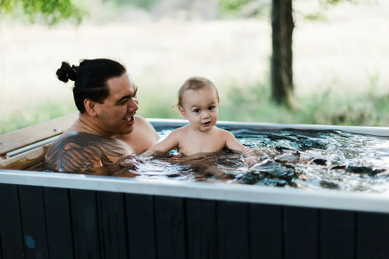 Kamalu with his child sitting together in an outdoor tub, with a tree and grassy field behind them