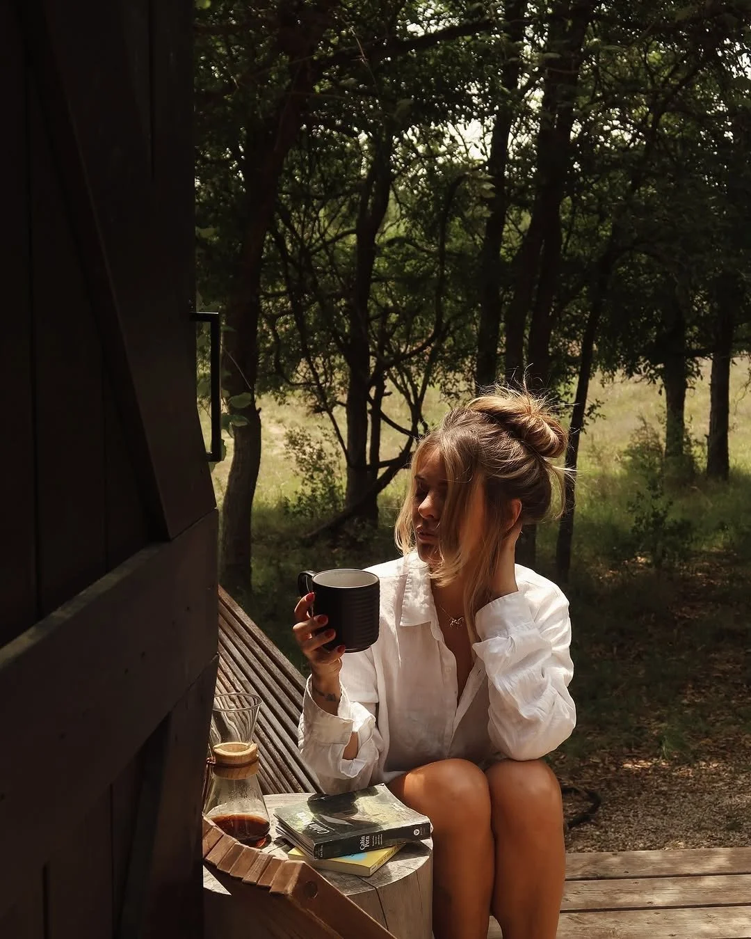 A guest in a white shirt sits on a wooden deck holding a black mug beside stacked books and a glass bottle.
