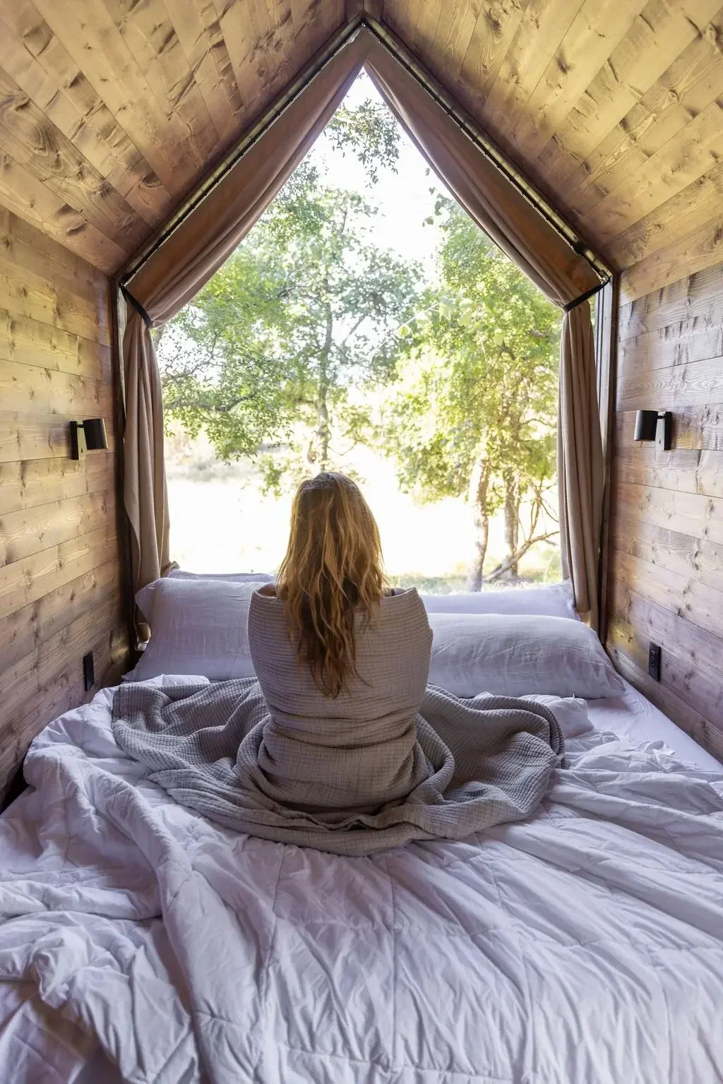 A guest sits on a bed inside a wood-paneled cabin, facing a large triangular window overlooking trees.