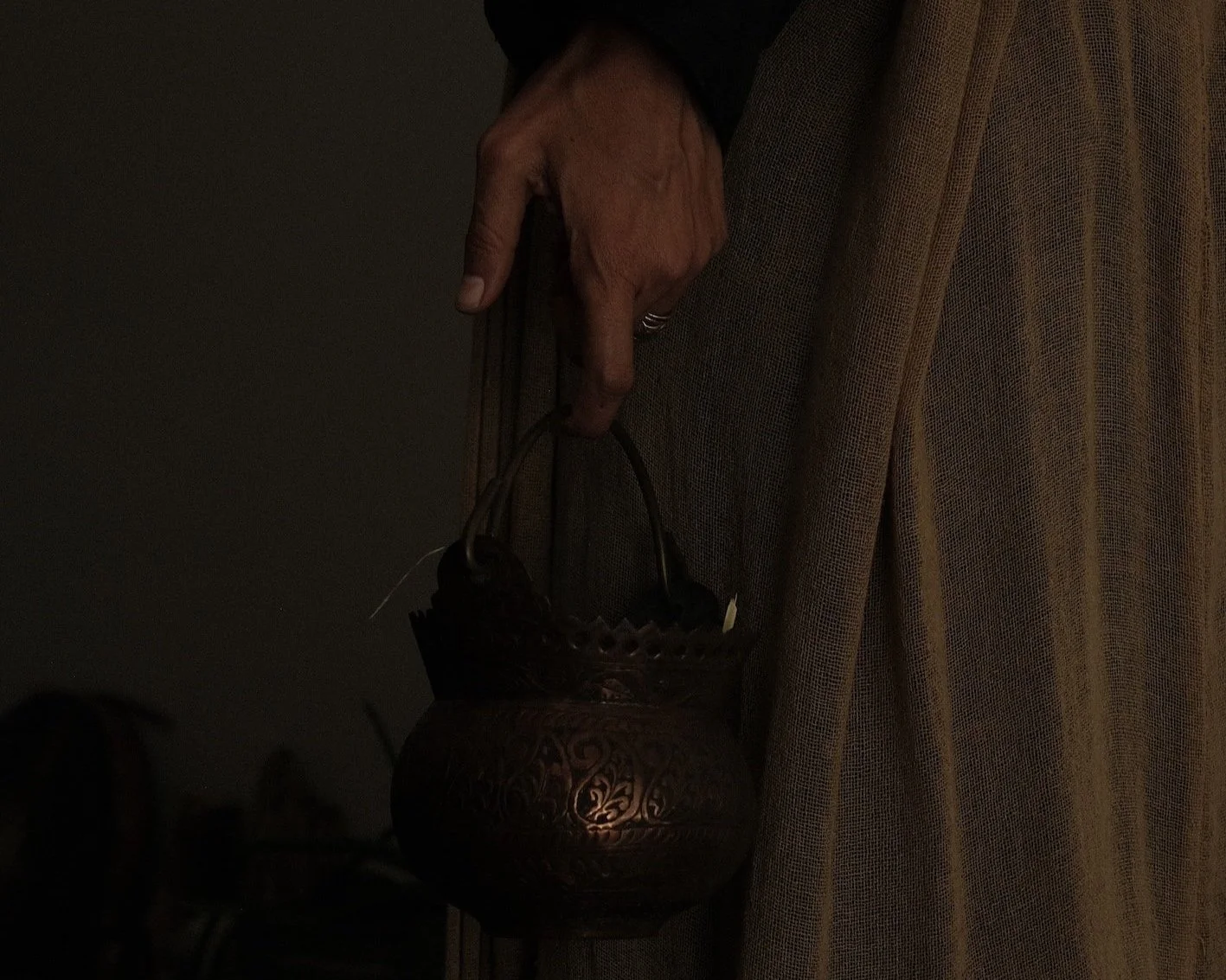 A person's hand holding an ornate metal lantern with intricate patterns, partially covered by a long, light-colored textured fabric or curtain.