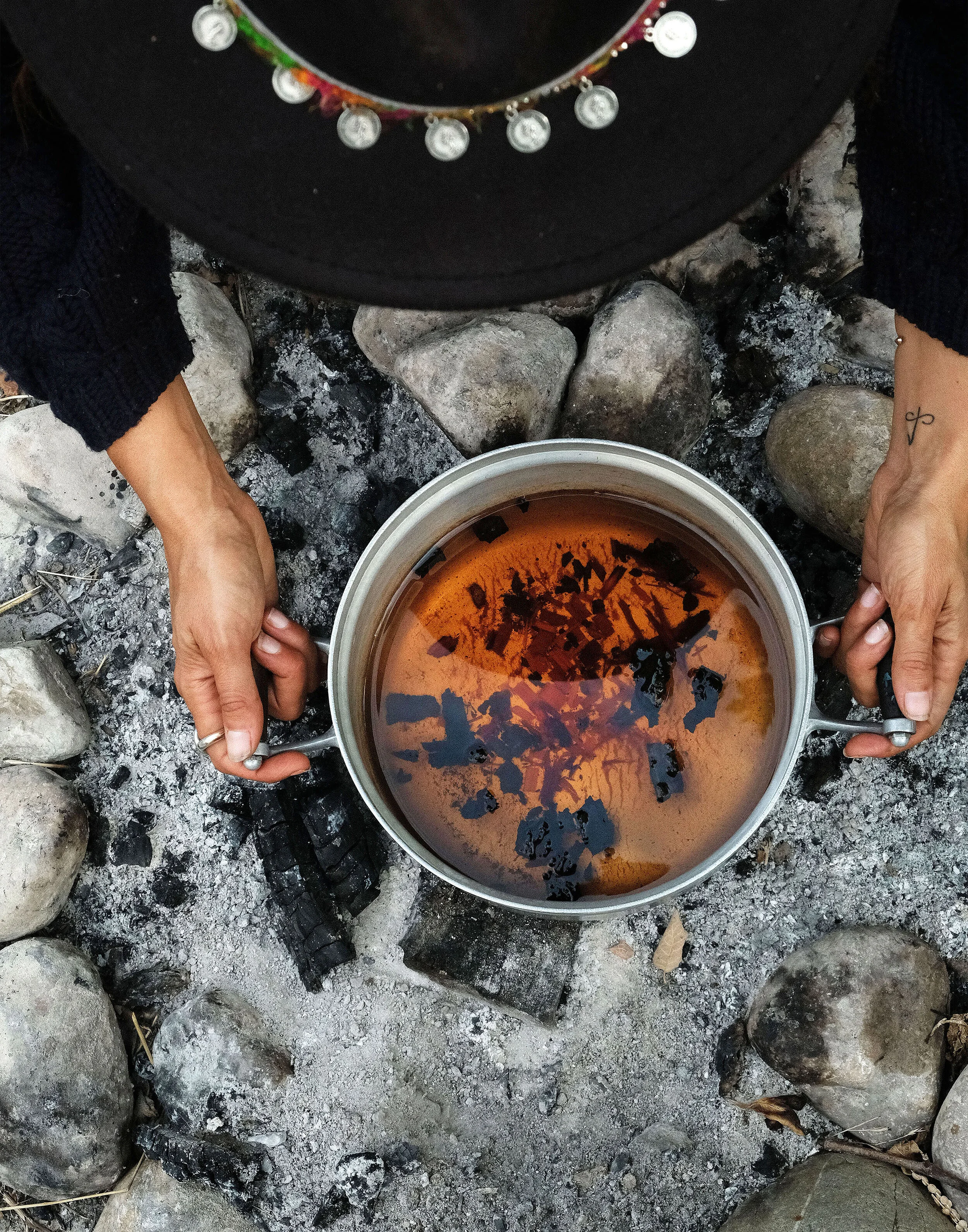 Overhead view of a person's hands holding a metal pot filled with steaming hot tea over an outdoor fire pit made of stones, with ashes and charcoal.