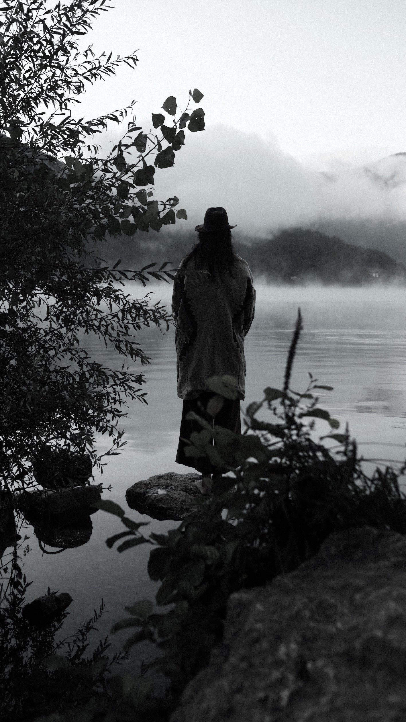 severine perron Poétesse, artiste, autrice. A person stands by a lake, surrounded by foliage, with misty mountains in the background.