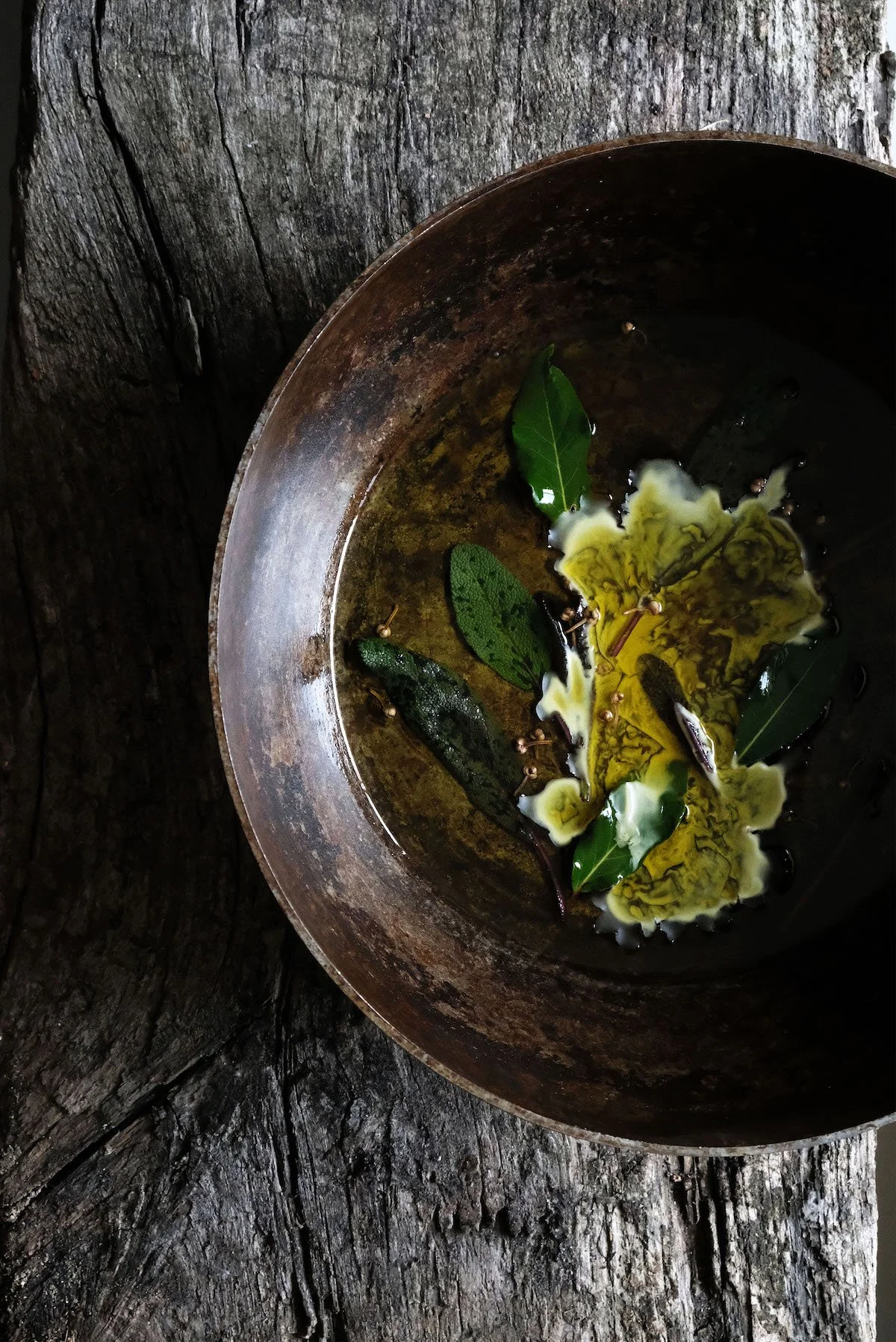 Close-up of a rusty metal bowl with green leaves and a yellowish liquid inside, placed on a rough wooden surface.
