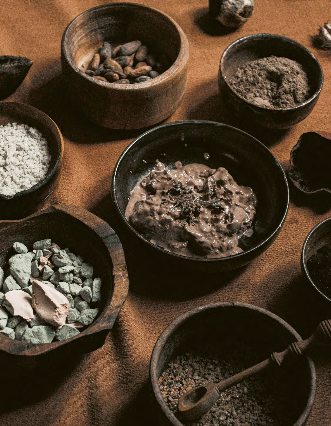 Dark wooden bowls and boxes filled with various textured ingredients such as rocks, powder, seeds, and a chocolate-based mixture on an orange-brown cloth surface.